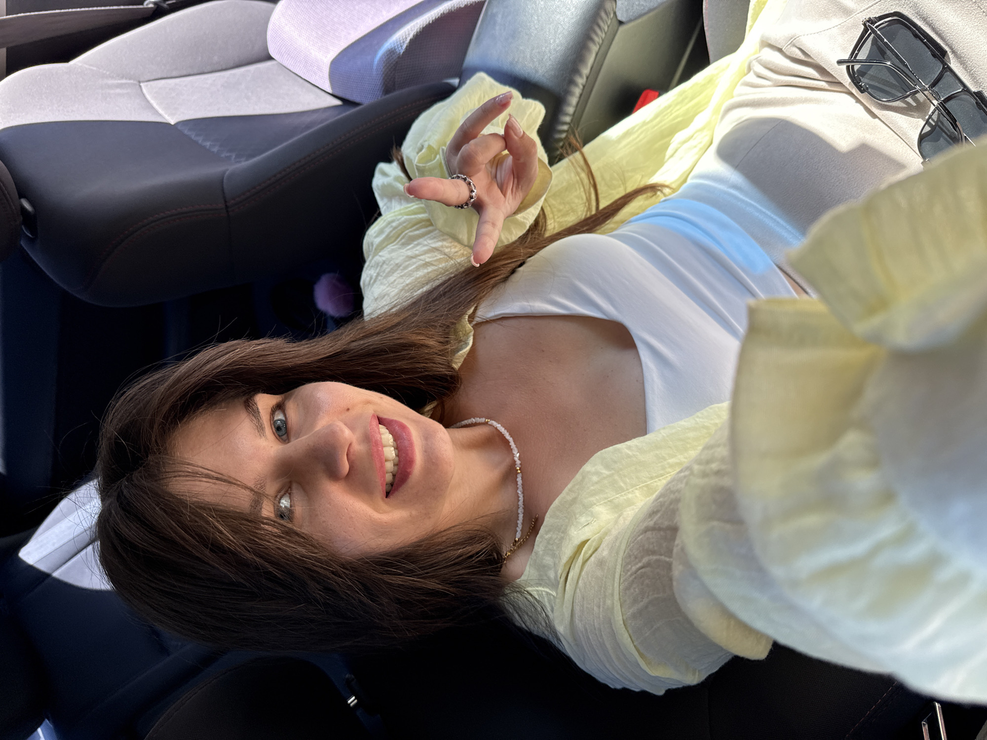 A woman with long brown hair and blue eyes smiles at the camera, her head slightly tilted. She is wearing a white tank top and a light yellow, open-front cardigan. A necklace made of small beads adorns her neck. Her left hand is raised, forming a peace sign, and she wears a ring on her middle finger. She is seated in the passenger seat of a car. Sunglasses are visible on her lap. The interior of the car, with dark upholstery and a lighter grey seat, is partially visible.