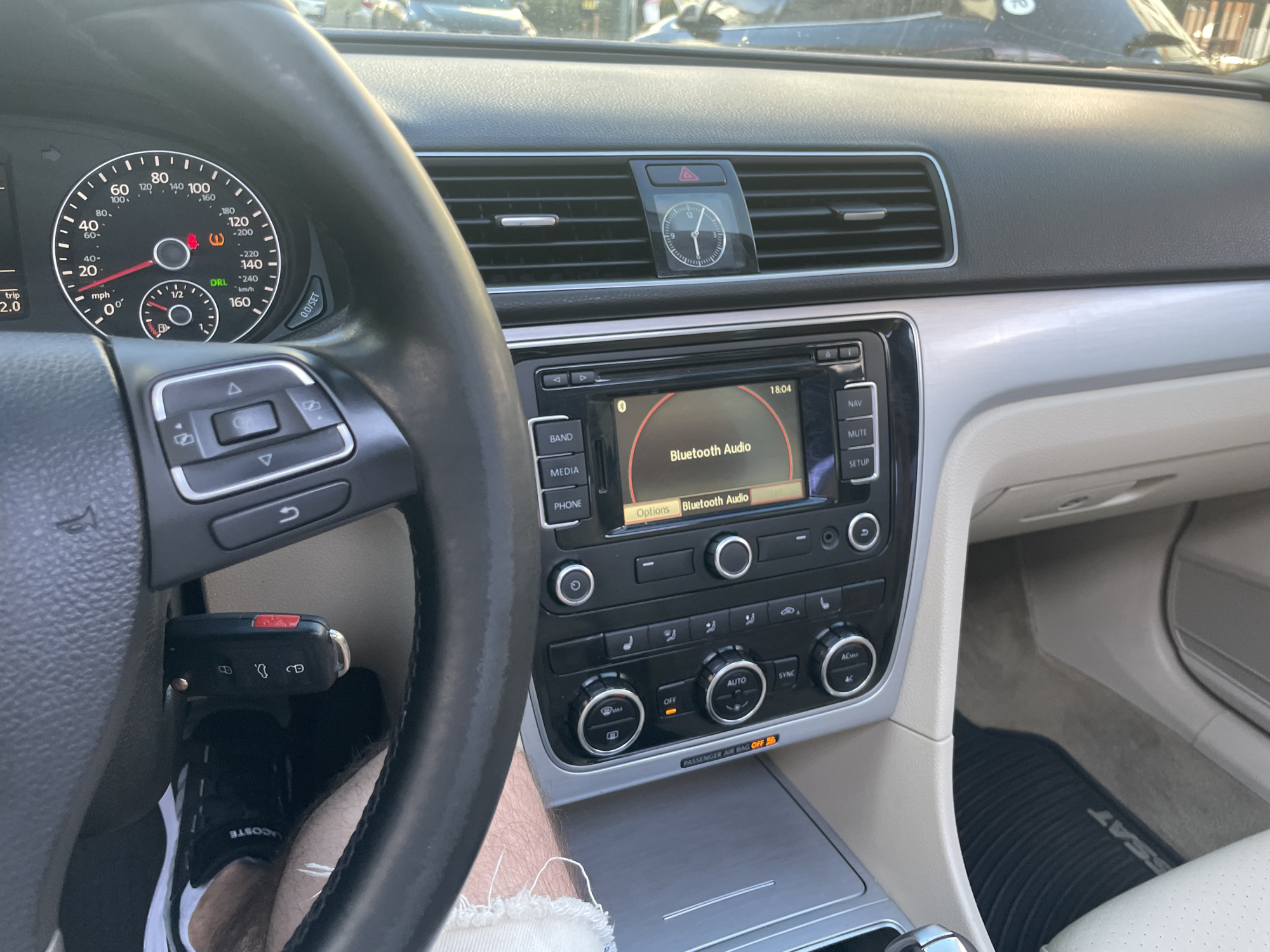 The image shows the interior of a Volkswagen car. A key fob is inserted into the ignition. The car's dashboard displays speed, RPM, and various warning lights, including a tire pressure warning. The infotainment system is active, showing "Bluetooth Audio" and the time as 18:04. A clock is mounted above the central air vents. The driver's seat is partially visible, along with the steering wheel. The overall scene suggests the car is parked, possibly in a residential area or a parking lot, given the background visible through the windshield. The image does not provide any specific information about the city of Kyiv, Ukraine.