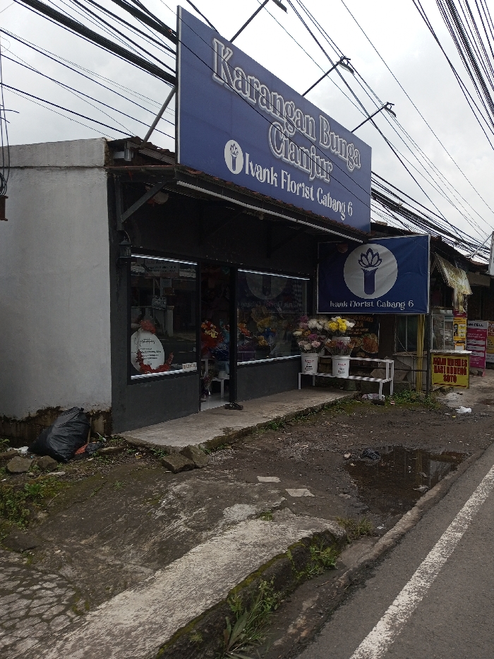 The image depicts the exterior of "Karangan Bunga Cianjur Ivank Florist Cabang 6," a florist shop located in the city of Chendenbji, Bhutan, under an overcast sky. The single-story building features a prominent blue rectangular sign above its facade, displaying the shop's name and a white lotus-like logo in white text. A smaller, square blue sign with the same branding hangs to the right. The shop entrance consists of two glass display windows showcasing various colorful flower arrangements, with a central doorway where a pair of dark slippers are visible on the interior floor. Outside the right display window, a white metal shelf holds three white buckets filled with different cut flower bouquets. To the immediate left of the building, a black trash bag rests on the ground near the base of the structure. The ground in the foreground comprises a partially cracked concrete sidewalk that transitions into an uneven dirt path, featuring a significant puddle of water on the right. A paved road with a white lane marking is visible on the far right. Overhead, a dense network of black utility wires traverses the scene. Partial views of other structures and signage are discernible to the far right, including a yellow sign. No individuals are visible.