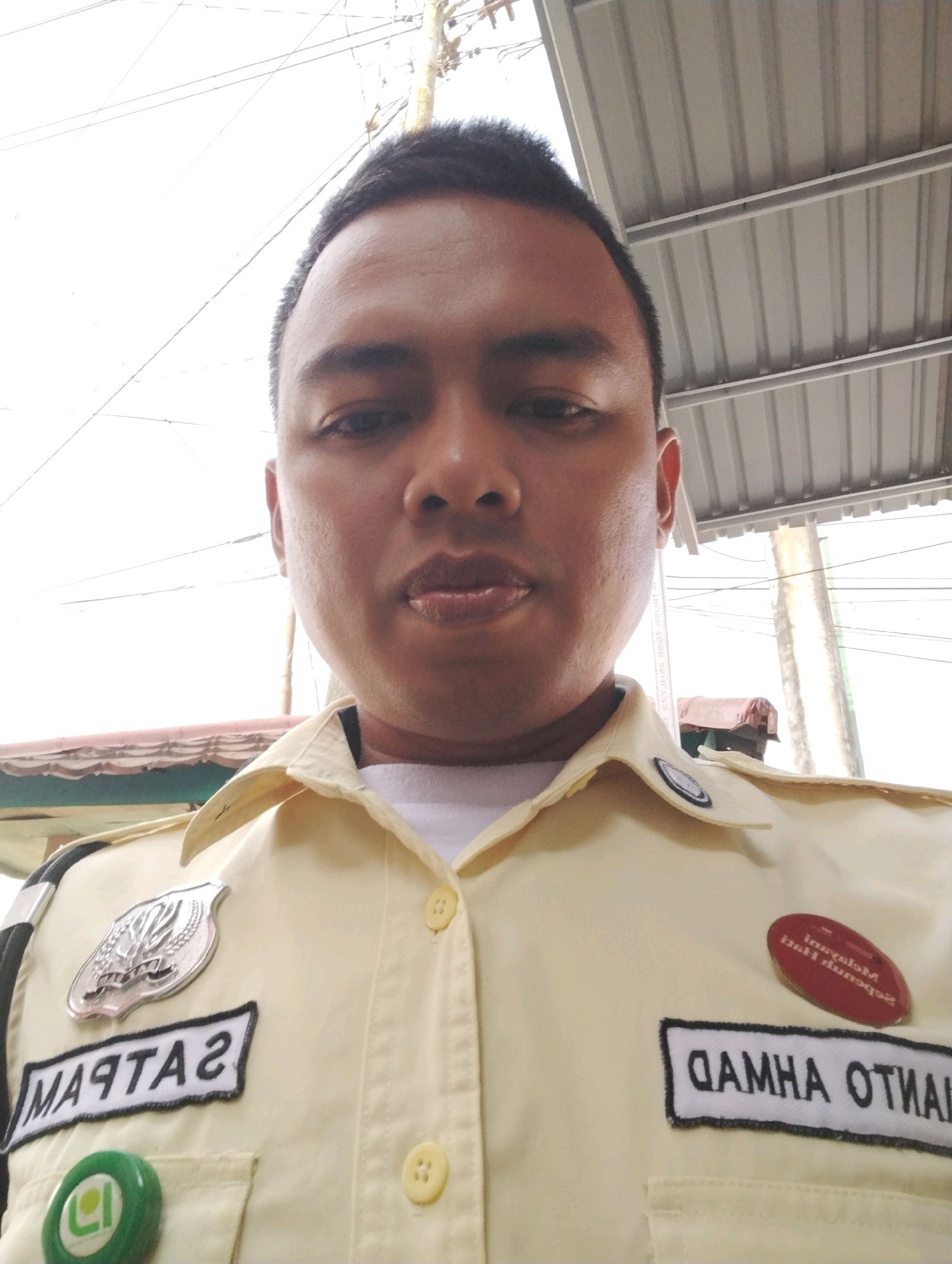 A close-up, waist-up selfie of an adult male wearing a light yellow uniform shirt. The man's face is centered in the frame, with his head tilted slightly down. He has dark hair and is looking directly at the camera. The uniform shirt has several patches and buttons visible. On the left chest is a silver shield-shaped emblem and below it a black rectangular patch with white lettering that reads "MARTAS". There is also a green circular pin with a white logo. On the right chest, there is a red oval pin with white lettering that is partially obscured and difficult to read. Below that is another black rectangular patch with white lettering that reads "GAMHA OTHAI". The background is out of focus but appears to be an outdoor setting with a corrugated metal roof overhead and some wires. The overall impression is that the photo was taken casually, possibly during a break. The specific location of Kuala Samboja, Indonesia, is not evident from the visual cues in the image itself.