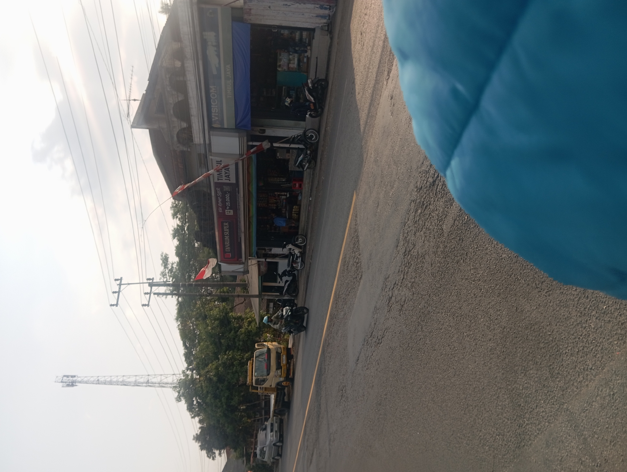 This is a medium shot taken from a low angle, looking across a road towards a commercial strip in Klakah, Indonesia. The foreground is dominated by the out-of-focus blue fabric of what appears to be a jacket or bag. The road itself is asphalt, with a yellow line dividing the lanes. In the middle ground, a person rides a dark-colored motorcycle. Parked on the side of the road are several other motorcycles, a yellow flatbed truck, and a silver van. Behind the vehicles, a row of shops is visible. One shop has a prominent "VISICOM" sign. Another shop has signage that includes "DJARUM SUPER" and "TIMUL JAYA". A red and white Indonesian flag is flying from a pole in front of one of the shops. In the background, a tall cell tower stands against a hazy sky, and lush green trees are visible. The overall impression is a bustling, everyday scene on a commercial street in Indonesia.