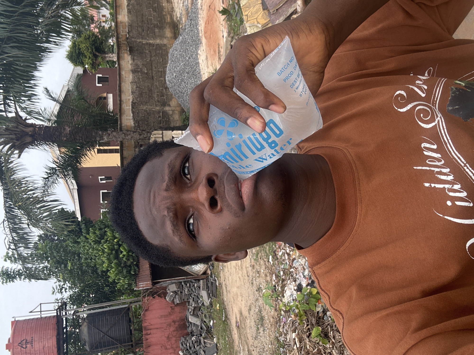A young man is holding a sachet of "miriugo" sachet water up to his lips, seemingly about to drink from it. He is wearing a brown long-sleeved shirt with white text. The photo appears to have been taken outside, in what looks like an urban area or a roadside in Orifite, Nigeria, indicated by the sachet water brand. In the background, there are buildings, palm trees, and some piles of rubble or construction materials. There's also what looks like a water tank. Litter is scattered on the ground near the subject.