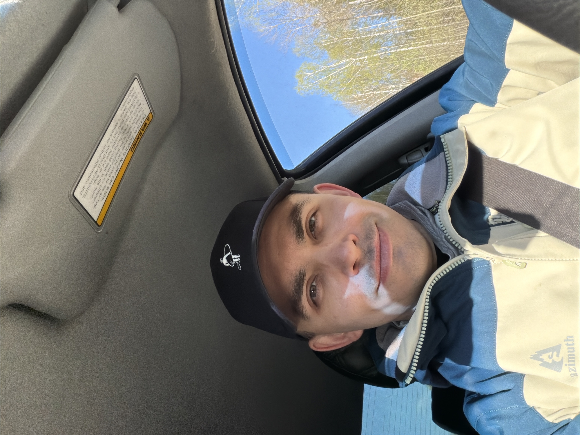 A man in a car is taking a selfie. He is wearing a black baseball cap with a white logo, a blue and white jacket, and a grey sweater. The man is looking at the camera with a slight smile. The car is grey and the sun visor is down on the driver's side. There is a sticker with a warning label on the sun visor. The sunroof is open and there are trees and a blue sky visible in the background. Based on the OCR content, the jacket has the word "azimuth" on it. The location is not discernible from the image.