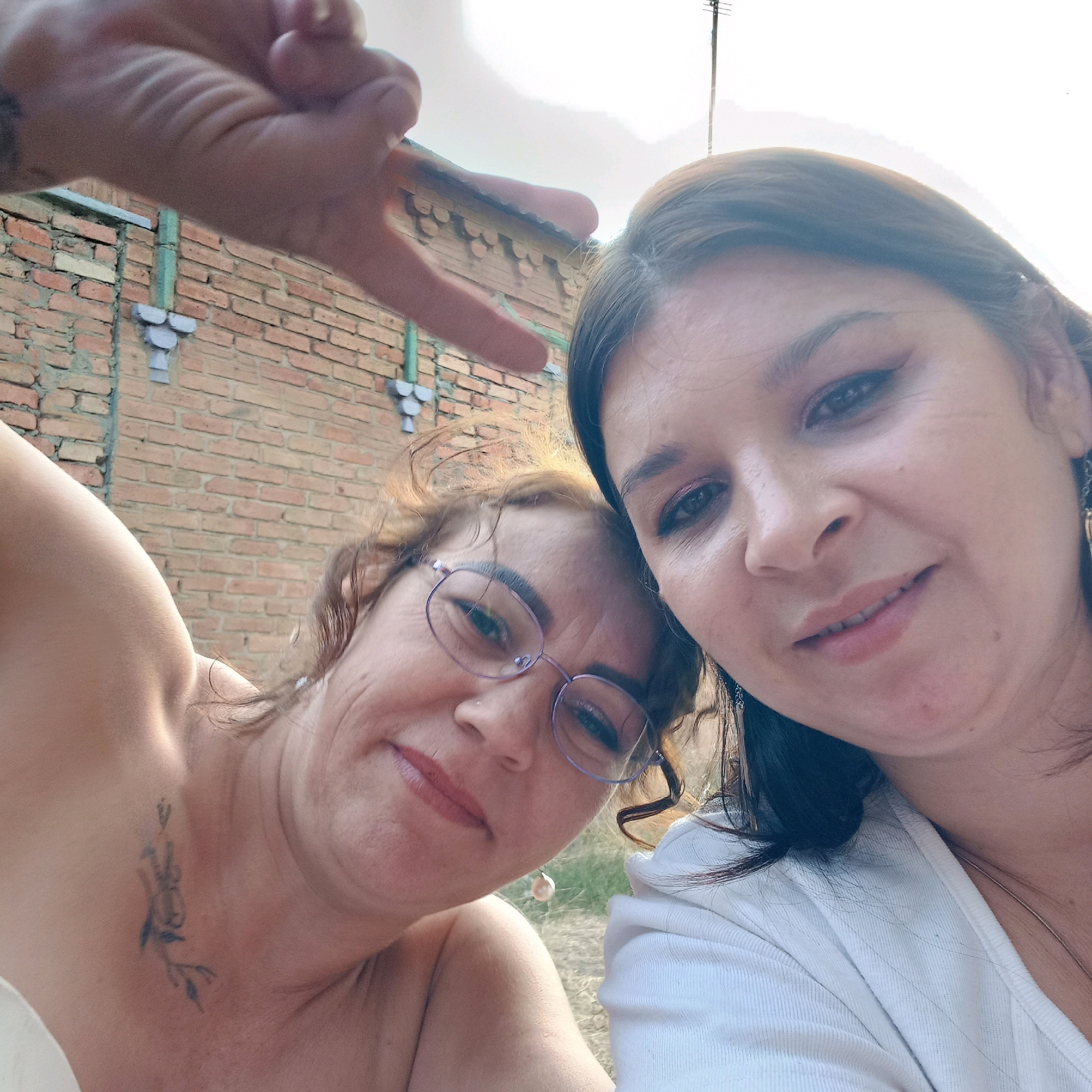 Two women are taking a selfie outdoors in front of a brick building. The woman on the left is closer to the camera and is wearing glasses and a pearl earring. She has a tattoo on her neck. The woman on the right is looking at the camera and is wearing a white shirt. A hand is visible in the upper left corner, making a peace sign. The background consists of a brick wall with some green and purple decorative elements, a utility pole, and a hazy sky. The image is a close-up shot, with the faces of the two women occupying most of the frame.