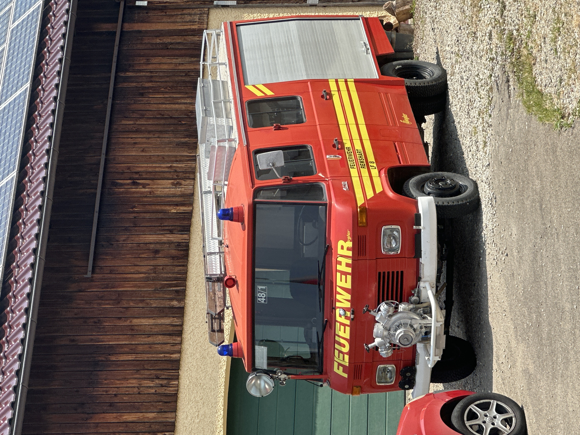 A red fire truck with yellow stripes is parked in front of a wooden building with solar panels on the roof. The fire truck has the word "FEUERWEHR" written on the front in yellow lettering. The truck also has two blue flashing lights on the roof and a spotlight on the front. The building appears to be a barn or garage, with a green door visible on the left side. The ground is covered in gravel and some dry grass. A portion of a red car is visible on the left side of the image. The scene appears to be in a rural or suburban area.