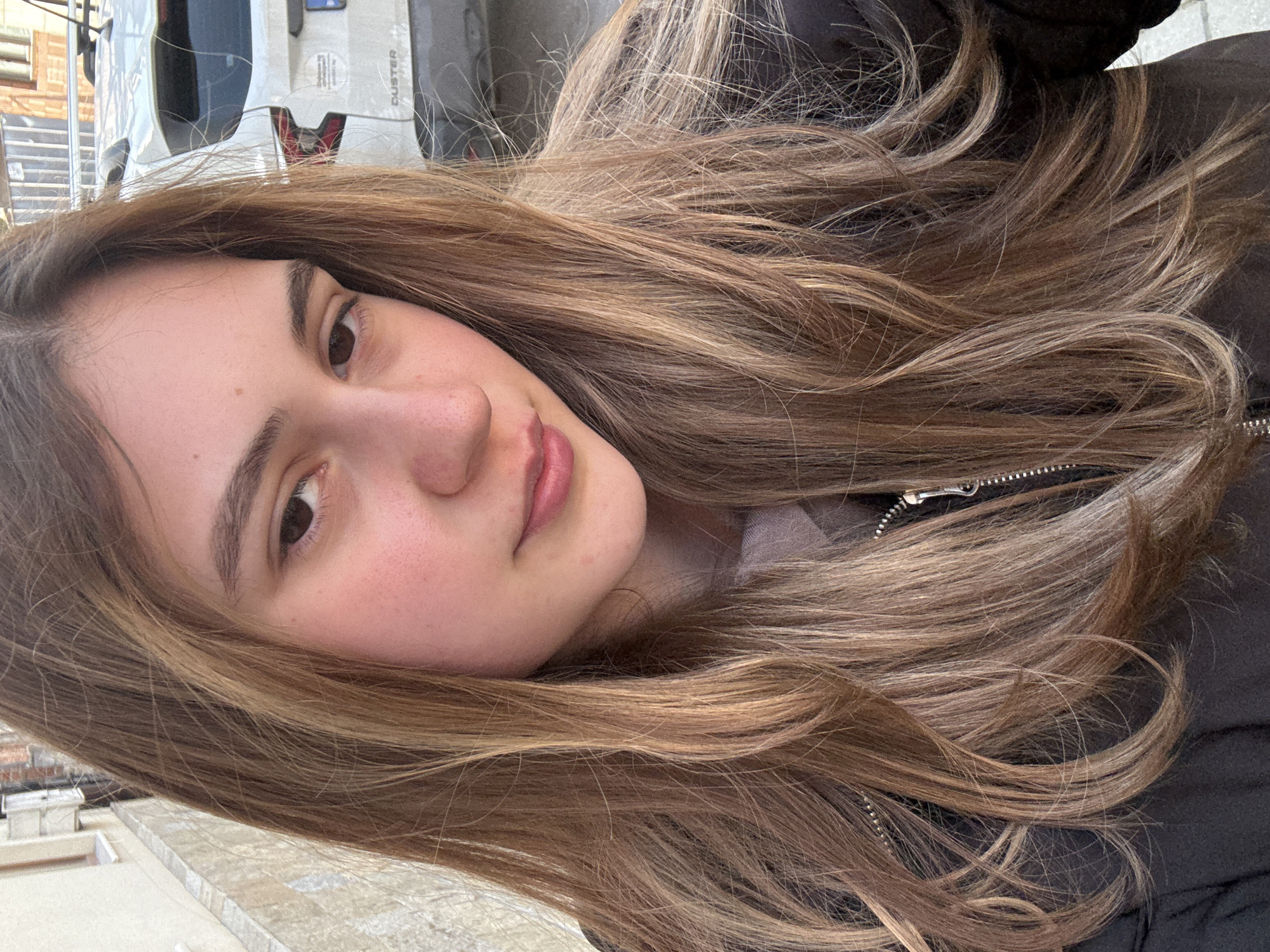 A young woman with long, wavy brown hair is the primary subject. She is wearing a black jacket with a zipper. The background includes a light-colored vehicle with the word "DUSTER" visible, suggesting a Dacia Duster model. A glimpse of a building and the street surface can be seen. The location is likely Panagyurishte, Bulgaria, given the prompt.