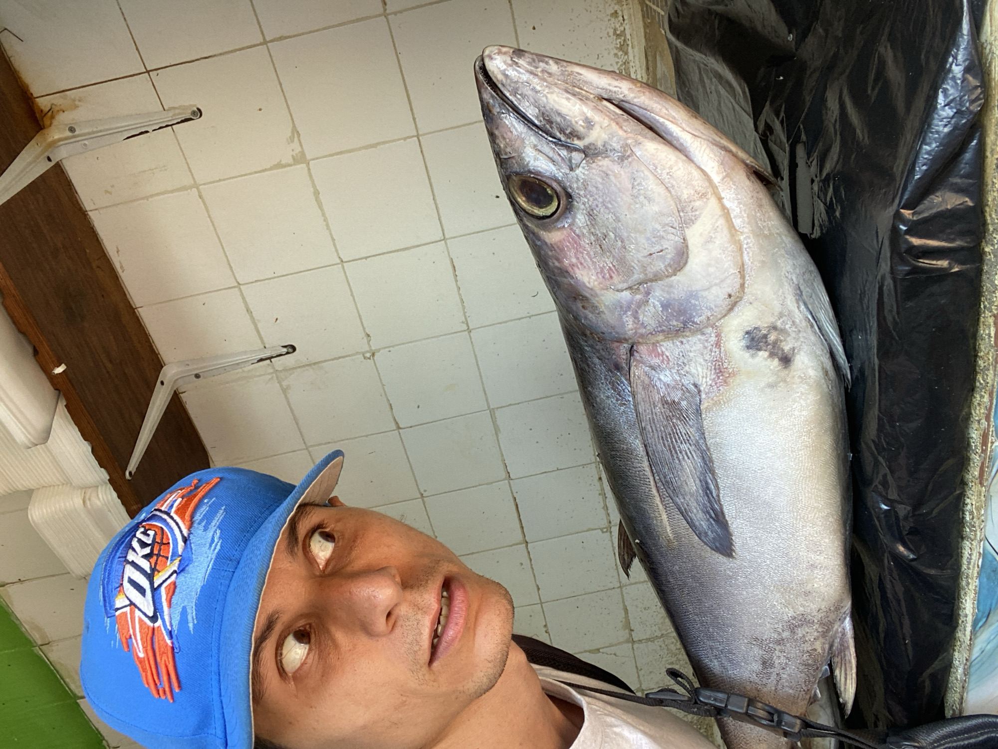 A man wearing a blue baseball cap with an "OKC" logo is posing for a selfie with a large, whole, silvery-grey fish. The fish is upright, positioned to the right of the man, and its head is near the top of the frame. The man's face is tilted upward toward the fish. The background consists of white tiled walls, brackets, and black material (possibly a plastic bag or liner). The scene appears to be indoors, potentially in a market or food preparation area. The photograph was taken in San José de Barlovento, Venezuela.
