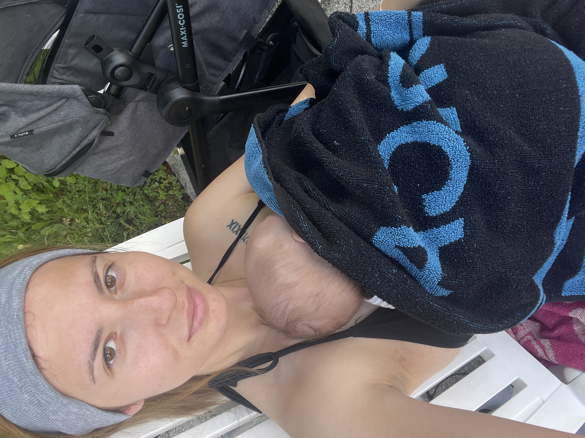 A young woman wearing a grey headband and a black bikini top sits on a white bench, holding a baby wrapped in a black and blue towel. The baby is facing away from the camera and is partially visible. To the right of the woman is a grey stroller with the brand name "MAXI-COSI" visible on it. The background shows green foliage, suggesting an outdoor setting, likely in the vicinity of a park or garden in Oberammergau, Germany. The overall lighting indicates it is daytime.
