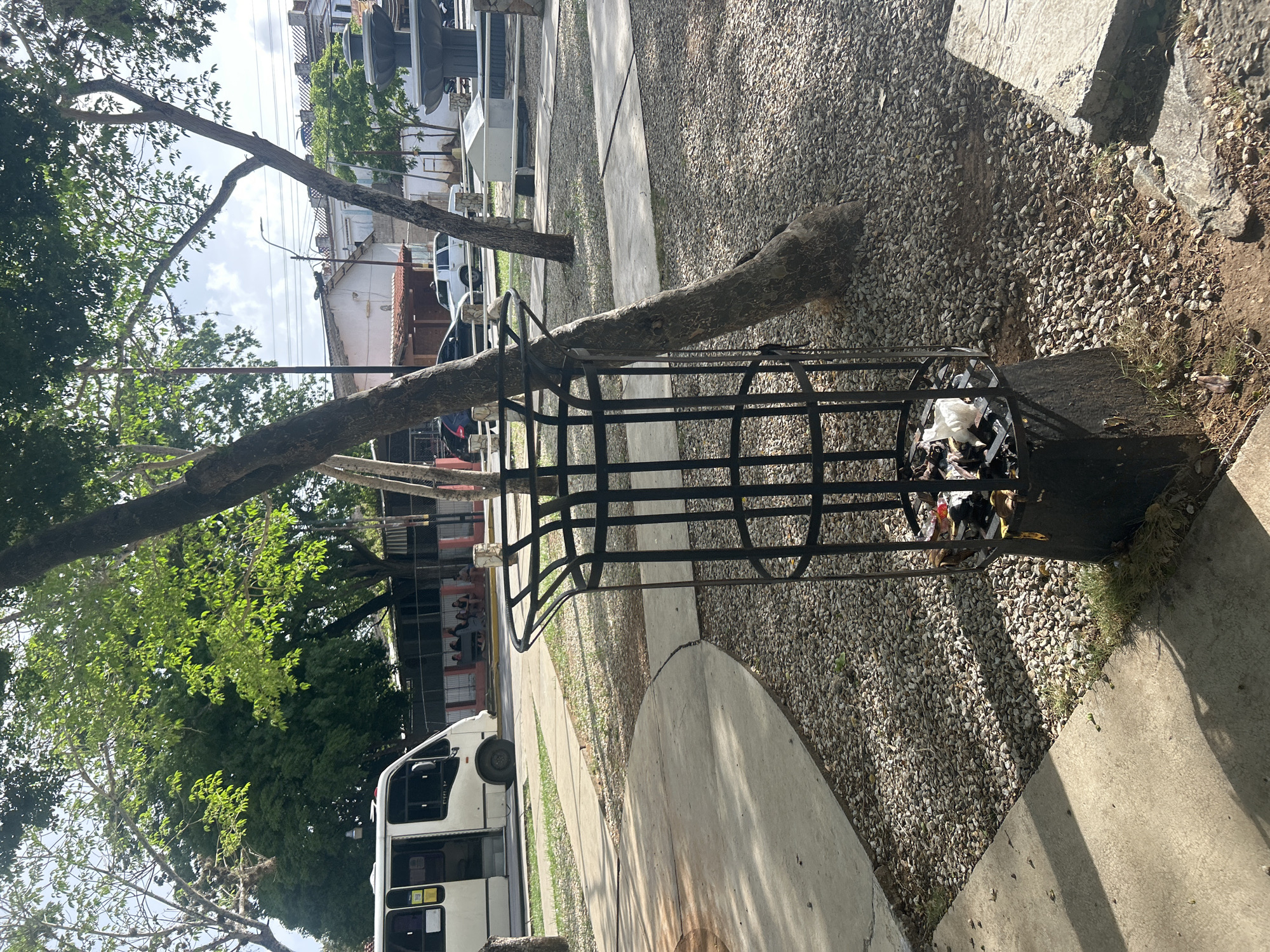 The image depicts an outdoor scene in the city of Santa Ana, Venezuela, likely a public space or park. In the foreground, a metal trash receptacle, partially filled with refuse, is situated on a gravel-covered ground. A concrete pathway curves around the receptacle and extends into the background. To the left, a white bus is parked on a paved area, with its door open. In the mid-ground, several people are seated on a bench near a fence. Further in the background, buildings and trees are visible, suggesting an urban or semi-urban setting. A water fountain can be seen on the right side of the image. The overall environment appears to be well-lit by natural sunlight, casting shadows on the ground.
