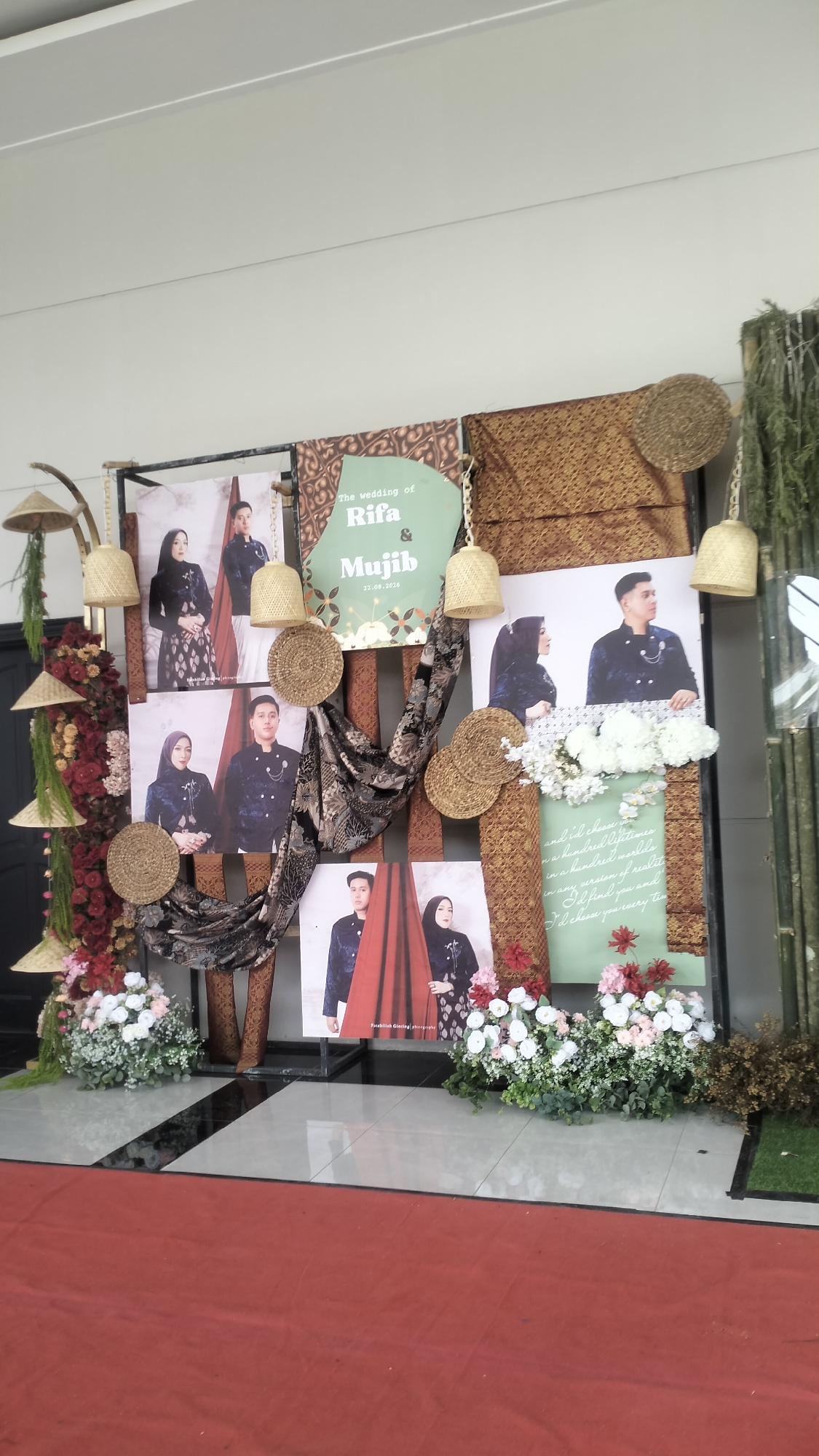 The image depicts a wedding photo display at the Hermes Palace Hotel in Lhokseumawe, Aceh, Indonesia. The display features several large portraits of a couple in traditional wedding attire, interspersed with decorative fabrics and floral arrangements. Woven basket-like decorations and bamboo stalks frame the display. The floor is tiled, and a red carpet is visible in the foreground, suggesting an event entrance or pathway. The overall presentation is festive and elegant, typical of a wedding reception setup.