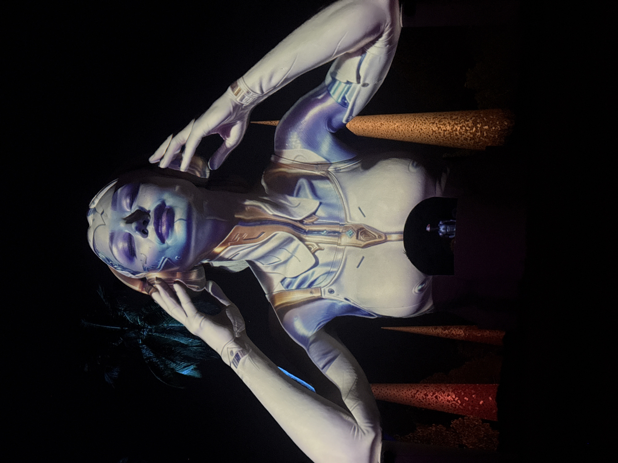 A projection of a female figure with blue and purple lighting on its face and body is displayed against a dark background. The figure wears a white bodysuit with gold accents. Its hands are raised to its head, and its eyes are closed. In the background, a palm tree is visible on the left, and several conical structures with textured surfaces are placed in front of the figure. A small projection of a standing figure is visible within an opening in the lower portion of the main figure's bodysuit. The overall impression is of a futuristic or sci-fi themed display. The location is unknown.