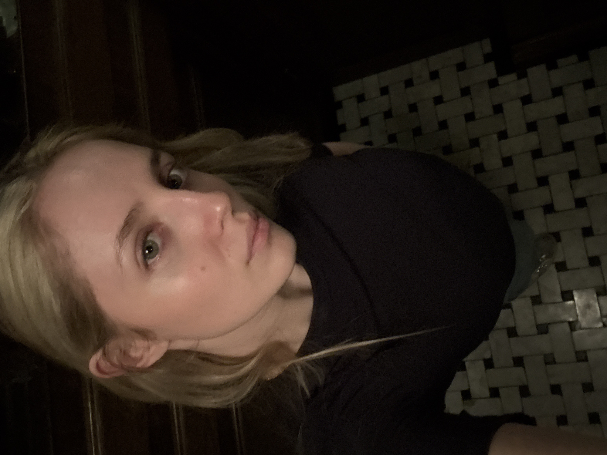 A young woman with blonde hair and fair skin is pictured from a low angle. She is wearing a black shirt. The background consists of a dark wooden surface to the left and a black and white tiled floor to the right, suggestive of a bathroom or similar space. The location appears to be a building in Mexico City, Mexico, as indicated by the distinctive tiled floor, which is a common architectural feature in many establishments throughout the city. The overall lighting is dim.
