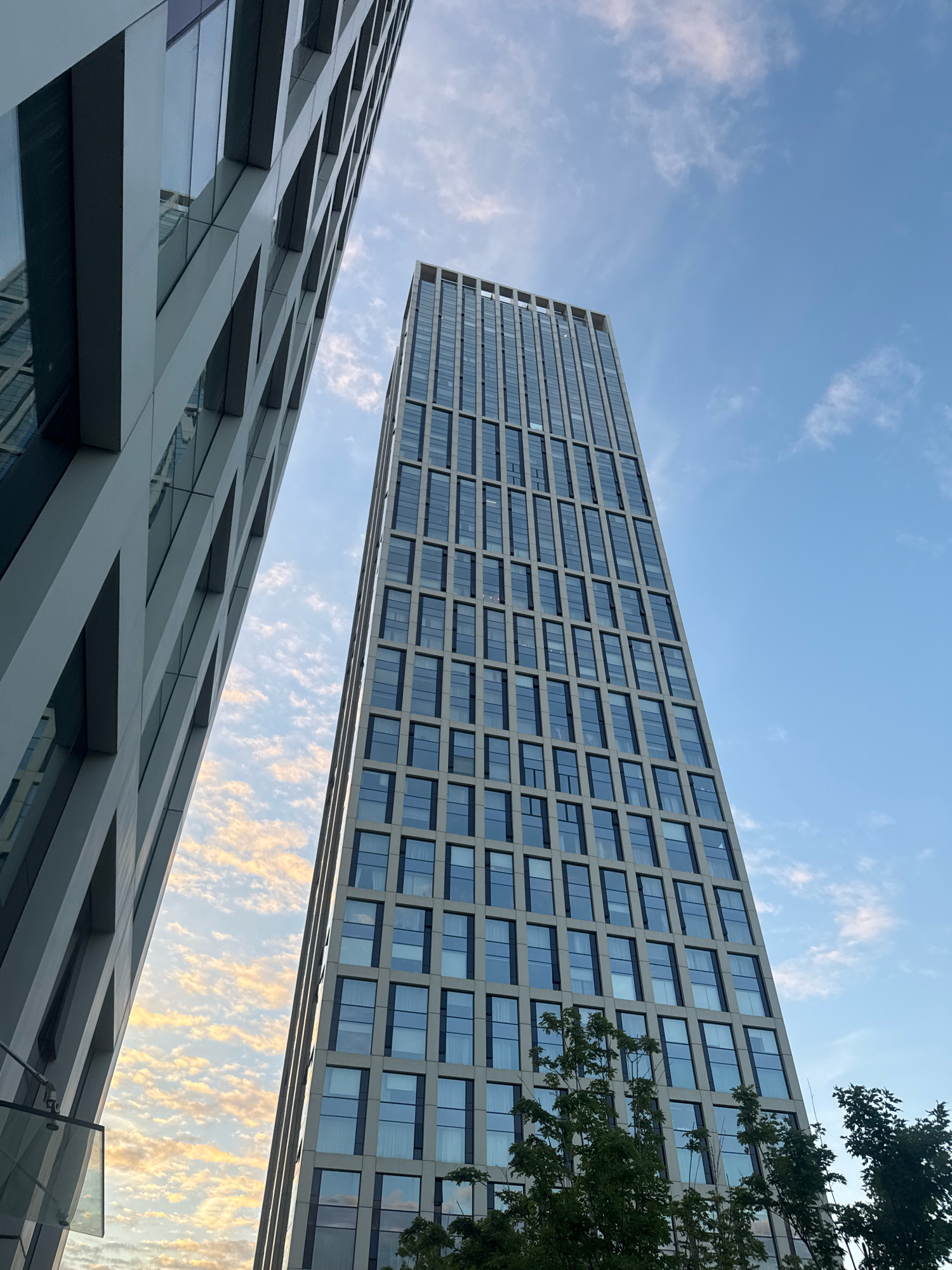 Two modern skyscrapers are viewed from a low angle against a partly cloudy blue sky. The skyscraper on the left is closer and appears to be under construction or has an unfinished facade with geometric patterns. The skyscraper on the right is taller and more distant, with a grid of windows. The sky is partly filled with wispy clouds, some of which are illuminated with a warm, golden light, suggesting it might be sunrise or sunset. Trees are visible in the foreground at the bottom of the frame. The image does not depict any people or specific actions. The architecture suggests an urban setting, likely a business district or a newly developed area of a city.