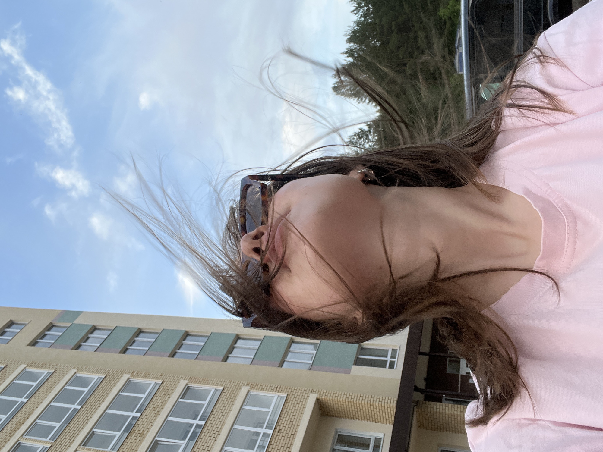 A person with brown hair and sunglasses, wearing a pink shirt, is photographed from a low angle with their hair blowing in the wind. Behind the person is a light brown building with multiple rows of rectangular windows with white frames. A blue sky with scattered clouds is visible in the upper portion of the frame. The picture appears to be taken in the city of Chaikovsky, Russia.