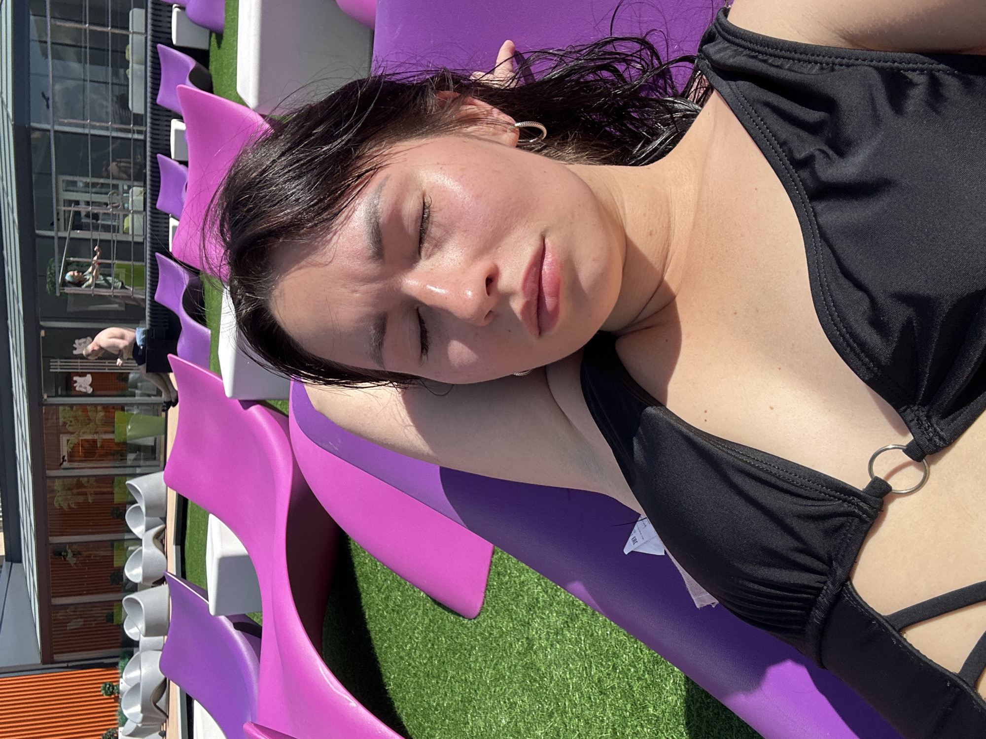 A young woman with dark hair and closed eyes lies on a purple chaise lounge. She is wearing a black swimsuit with a ring detail and hoop earrings. Her skin is slightly flushed, and there is sweat on her forehead. The chaise lounge is on a patch of artificial turf, with other colorful chaise lounges and white seating visible in the background. A building with large windows and orange striped panels is behind the seating area. The scene appears to be an outdoor recreational space, possibly a hotel or resort. There is no indication of the location being Saint Petersburg, Russia.