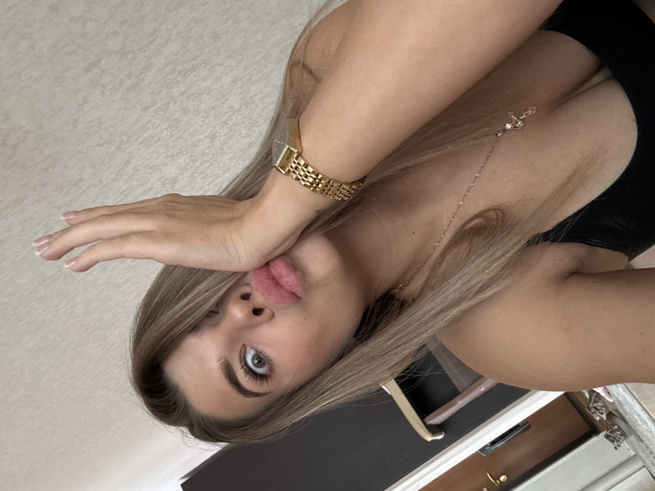 A female individual is depicted in a close-up, upward-angle indoor selfie. Her light brown hair is parted on the left, partially obscuring her right eye and forehead. Her visible left eye is wide open and directed towards the camera. Her lips are pursed forward in a pouting gesture. Her left hand is raised, palm inward with fingers slightly splayed, pressed against the left side of her face. She is wearing a dark-colored, low-cut tank top. A gold-toned rectangular wristwatch with a link band is worn on her left wrist. A thin gold-toned chain necklace with a small cross pendant rests on her chest. The background features a light-colored wall with a subtle texture. To the left, a dark vertical surface is partially visible, alongside a brown wooden door with a gold-toned handle. Indistinct light-colored objects are present on the floor to the lower left. A dark, possibly wooden, furniture piece is partially visible behind her left shoulder. The specific location within the city of Chaikovsky, Russia, cannot be determined from the visual information provided.