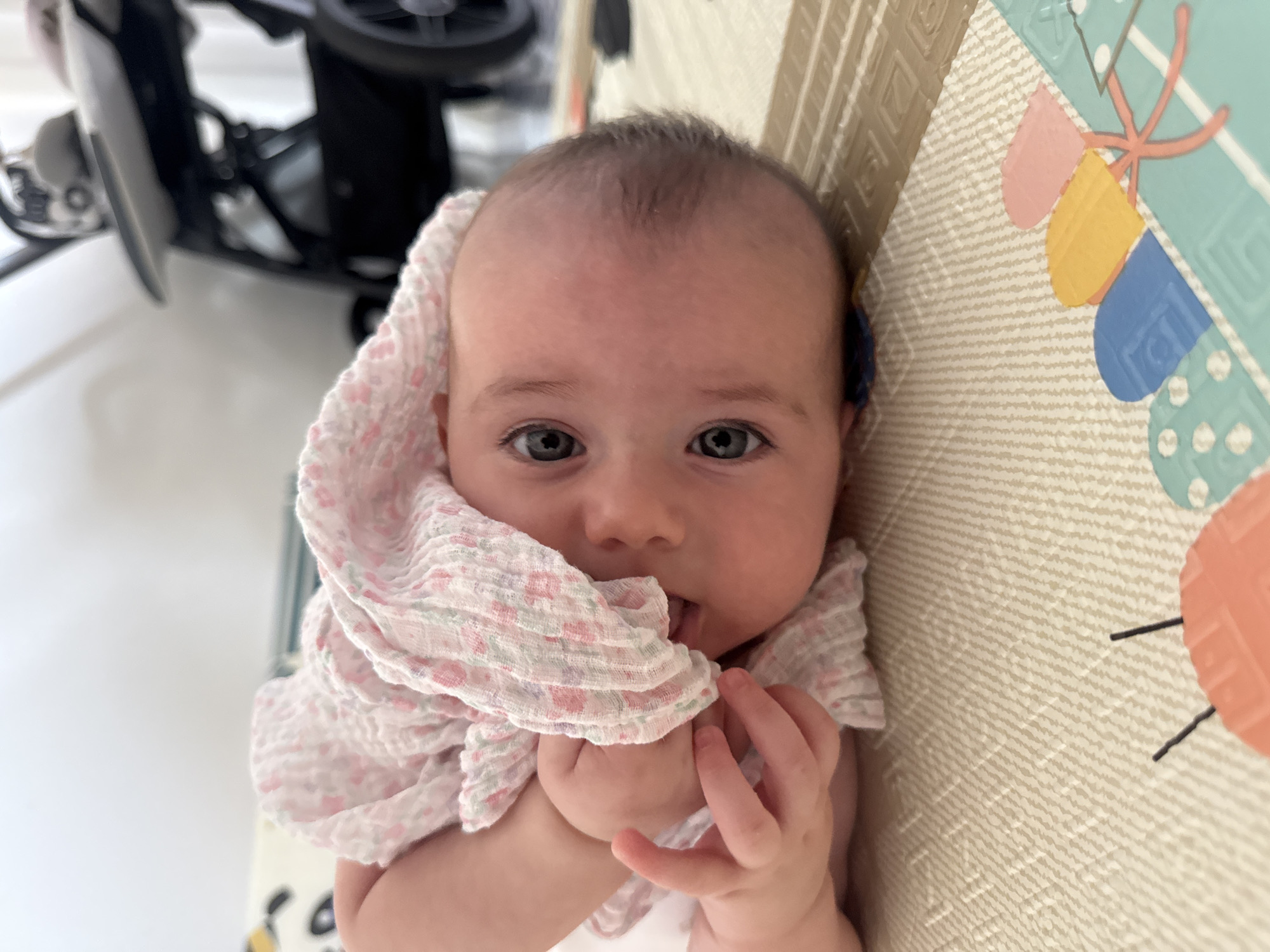 A baby is lying on its back on a play mat. The baby is wearing a pink floral print swaddle, part of which is draped over its head and face. The baby's left hand is raised and is touching its mouth. The baby has grey eyes and fair skin. In the background, a stroller is visible. The play mat has a textured surface with geometric patterns and colorful illustrations of objects. The scene is indoors, with a white wall in the background. There is no specific indication of the location being Isola delle Femmine, Italy.