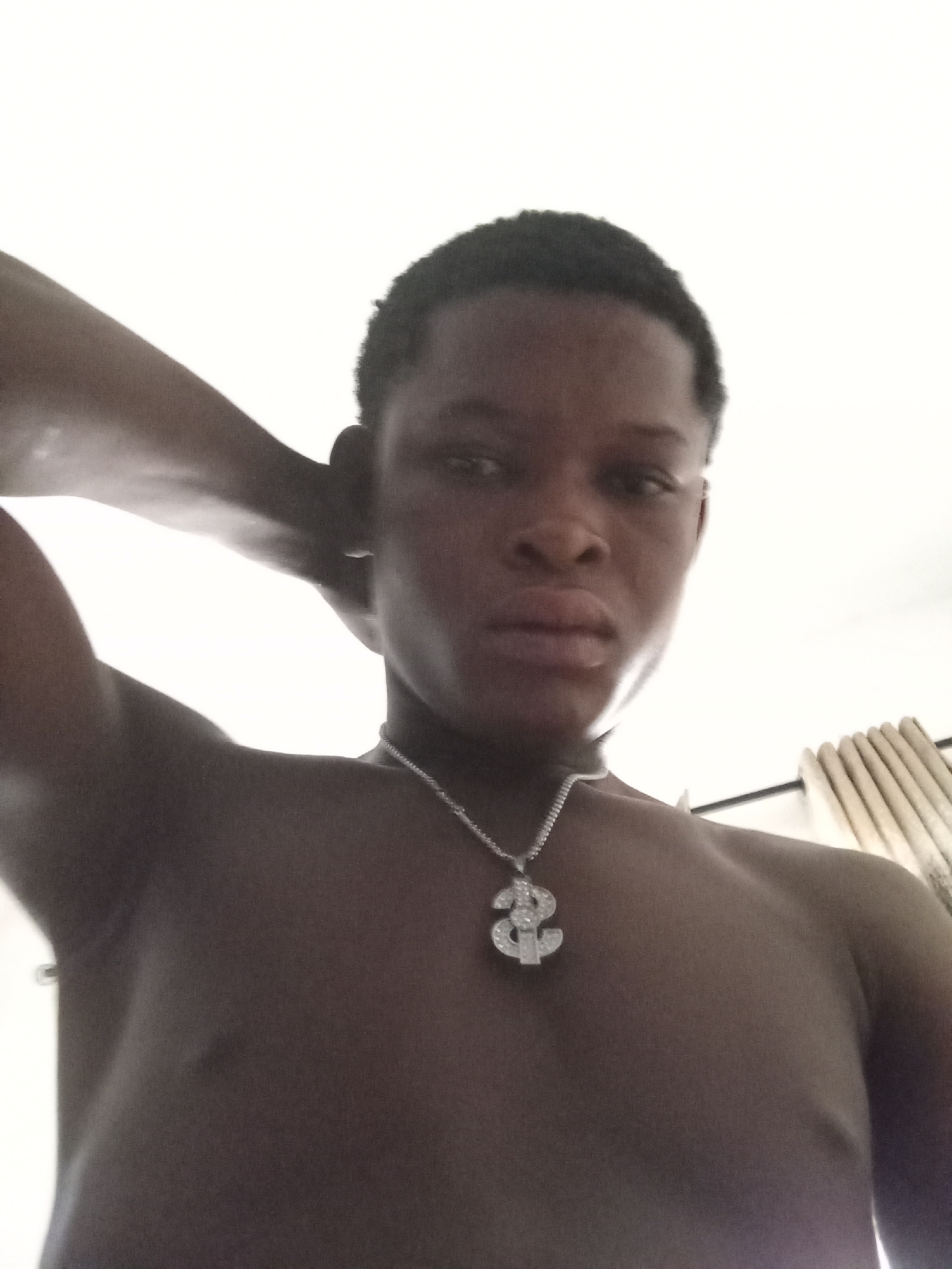 A low-angle, close-up, waist-up photograph shows a young Black male from Ado, Nigeria, from the chest up. The man has dark skin and short, dark hair. He is wearing a silver chain with a large, diamond-encrusted dollar sign pendant. His left arm is raised, with his hand resting behind his head. His right shoulder and arm are visible. The background is mostly white, with a hint of light brown curtains visible on the right side. The lighting suggests it may be daytime, possibly indoors with bright light from a window.