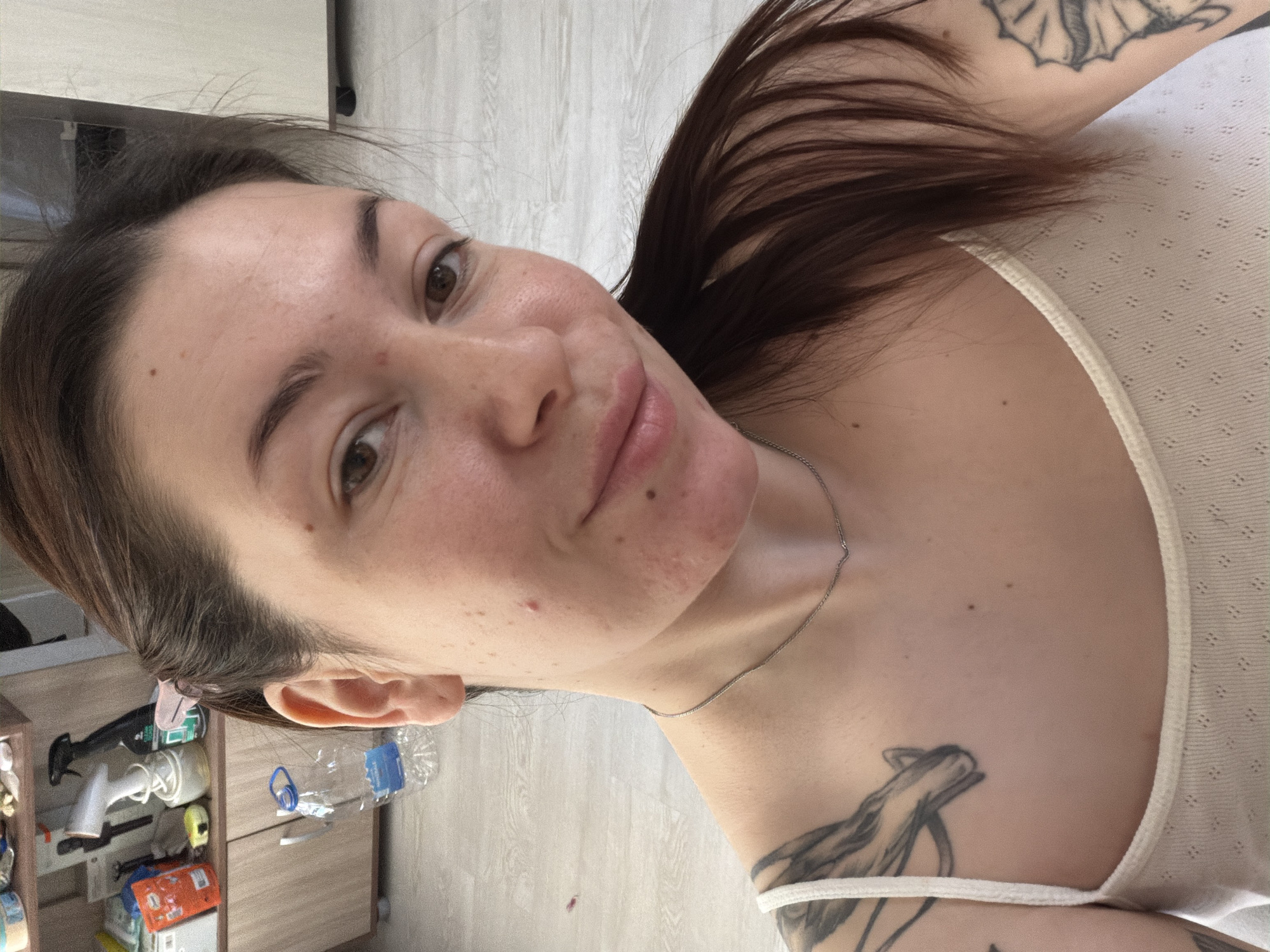 A woman is posing for a selfie indoors. She has brown hair, fair skin, and is wearing a light-colored tank top. The background includes a cabinet with various objects on it, including cleaning supplies and a water bottle. The location is not specified as Chaikovsky, Russia, and there is no way to verify this location from the image. The woman has tattoos on her chest and upper arm. She is wearing a thin silver necklace.
