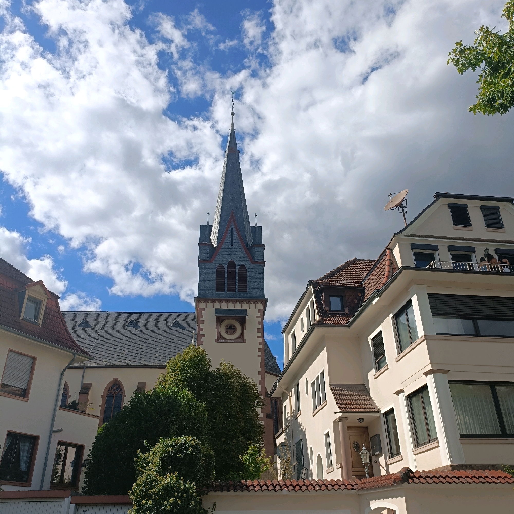 The image shows a view of the church tower of the Evangelical Church in Bad Kreuznach, Germany, from a street-level perspective. The church tower is tall and slender, with a grey, pointed roof and intricate stonework. It is flanked by residential buildings, which are typical of German architecture, with red-tiled roofs and light-colored facades. The sky is blue with scattered white clouds, and the sun is shining, casting shadows on the buildings. Trees and greenery are visible in the foreground and around the buildings, adding to the picturesque scene. The overall impression is one of a quiet, charming town with a rich history.