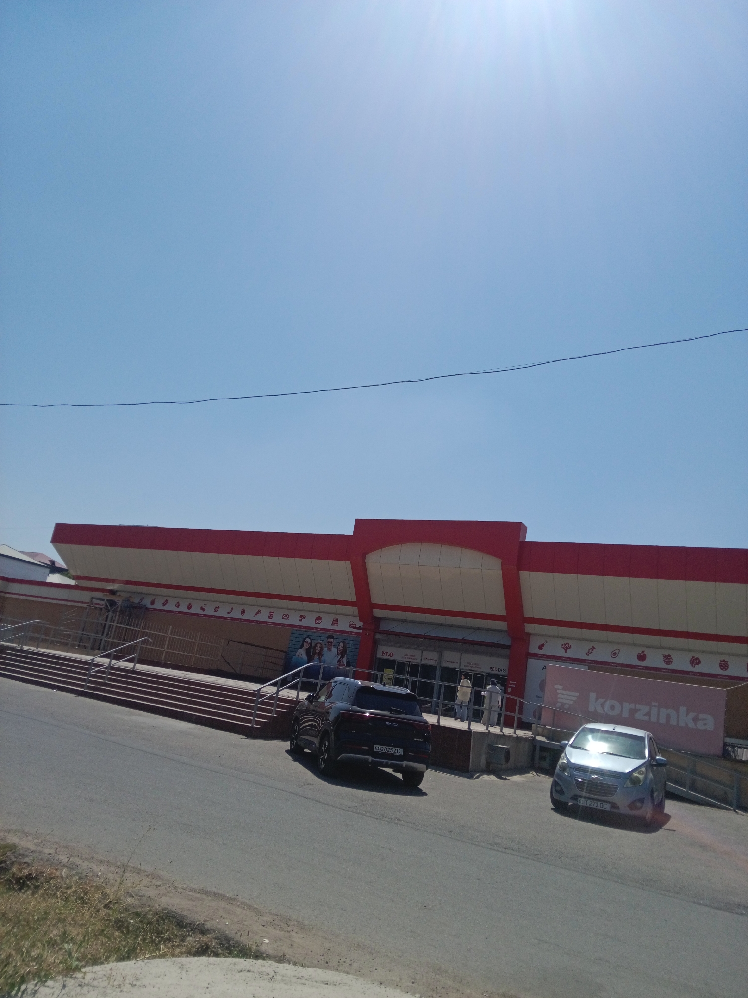 The image shows the exterior of a Korzinka supermarket in Tashkent, Uzbekistan. The building has a red and white facade with a prominent red roofline. Several cars are parked in front of the entrance, including a dark-colored SUV and a silver hatchback. A ramp leads up to the entrance, and there are steps on the left side of the building. A large sign with the Korzinka logo is visible on the right side of the building. The sky is clear and blue, indicating a sunny day.