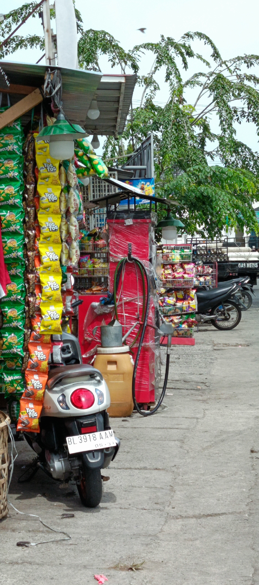 The image depicts a street-side vendor stall in Banda Aceh, Indonesia, likely in a busy area given the presence of traffic infrastructure and a truck. The stall is packed with various goods, predominantly snacks and drinks, displayed on shelves and hanging racks. Prominently featured are rows of brightly colored snack bags, including chips and biscuits, with some brands like "TOS TOS" visible. A silver scooter is parked in front of the stall, with its license plate reading "BL 3918 AAM". Behind the scooter, a large yellow plastic container sits next to a red structure covered in plastic wrap, possibly a refrigerator or display unit. A watering can is placed on top of the container. Further back, a black truck with the words "GAS MEDIS" printed on its side is parked, suggesting it might be a delivery vehicle for medical supplies or gas. The environment appears to be outdoors, with a concrete ground and trees with lush green foliage in the background, indicating a tropical climate. The sky is overcast, hinting at a possibly cloudy or rainy day. The overall scene captures a typical urban commercial setting in Banda Aceh, highlighting the daily economic activities of local vendors.
