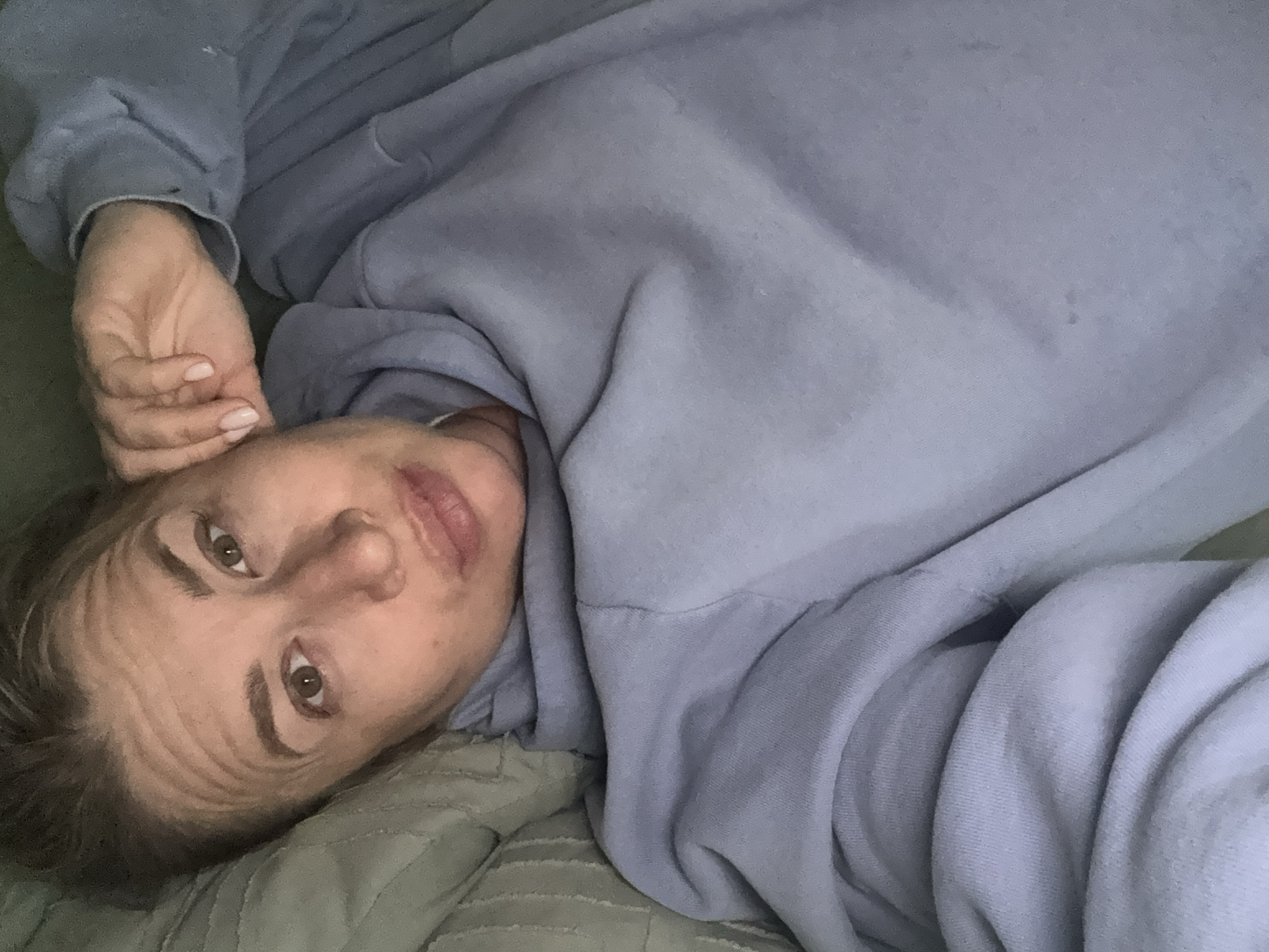 A person is lying down on a bed. The person is wearing a light blue sweatshirt. The background shows a green comforter with a quilted pattern. The photograph is taken at a slightly downward angle, with the person looking upwards towards the camera. There is no indication of Veliky Novgorod or any identifiable location within Russia.