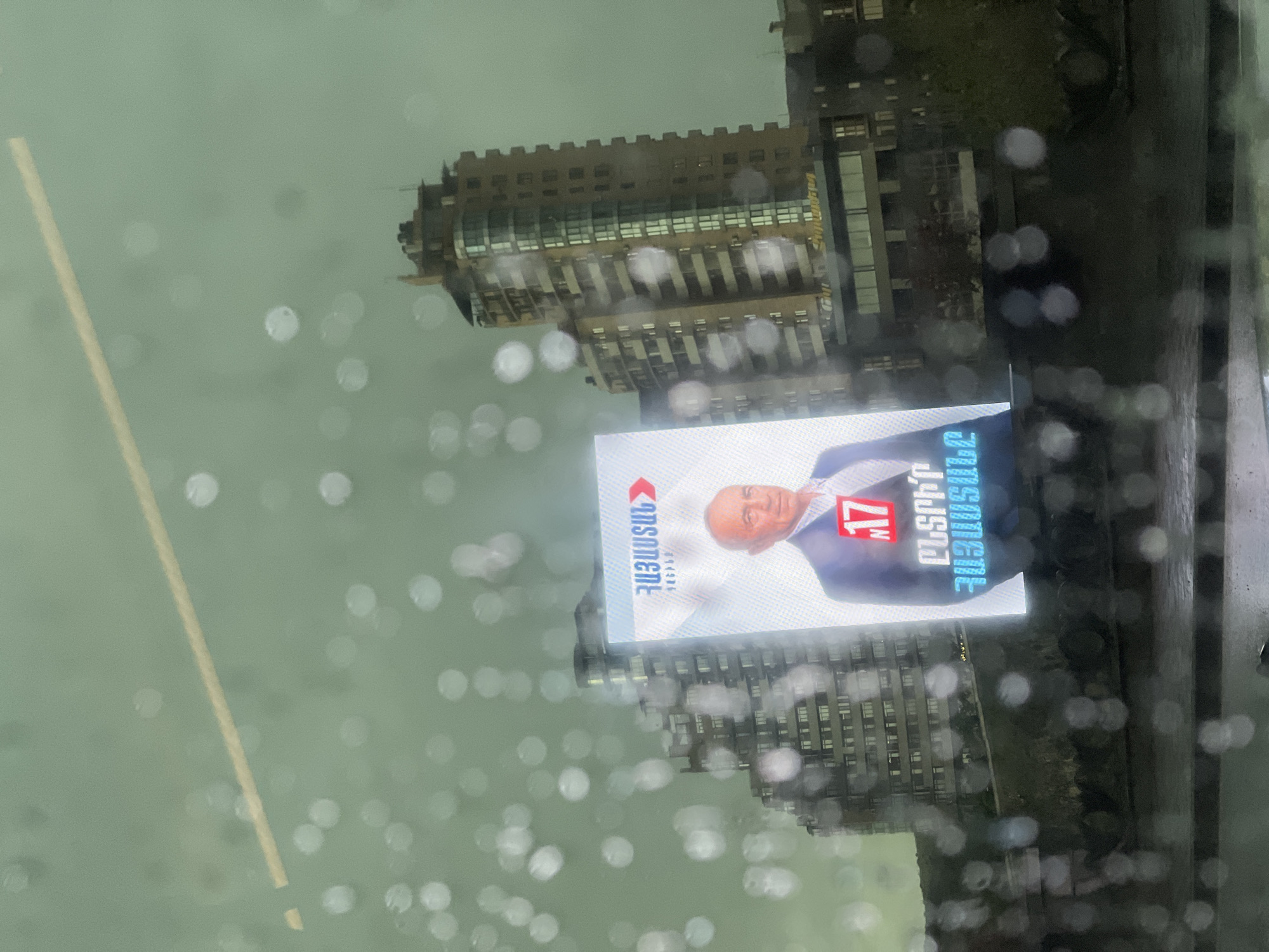 The image shows a cityscape scene, likely taken from inside a building or vehicle, as suggested by the raindrops on the glass. A prominent billboard is visible, displaying a political advertisement in Armenian, featuring a man in a suit and the number "17". The building style is consistent with architecture found in Yerevan, Armenia. The environment appears overcast or rainy, contributing to the blurred background. The buildings visible in the background are multi-story structures, typical of an urban setting.