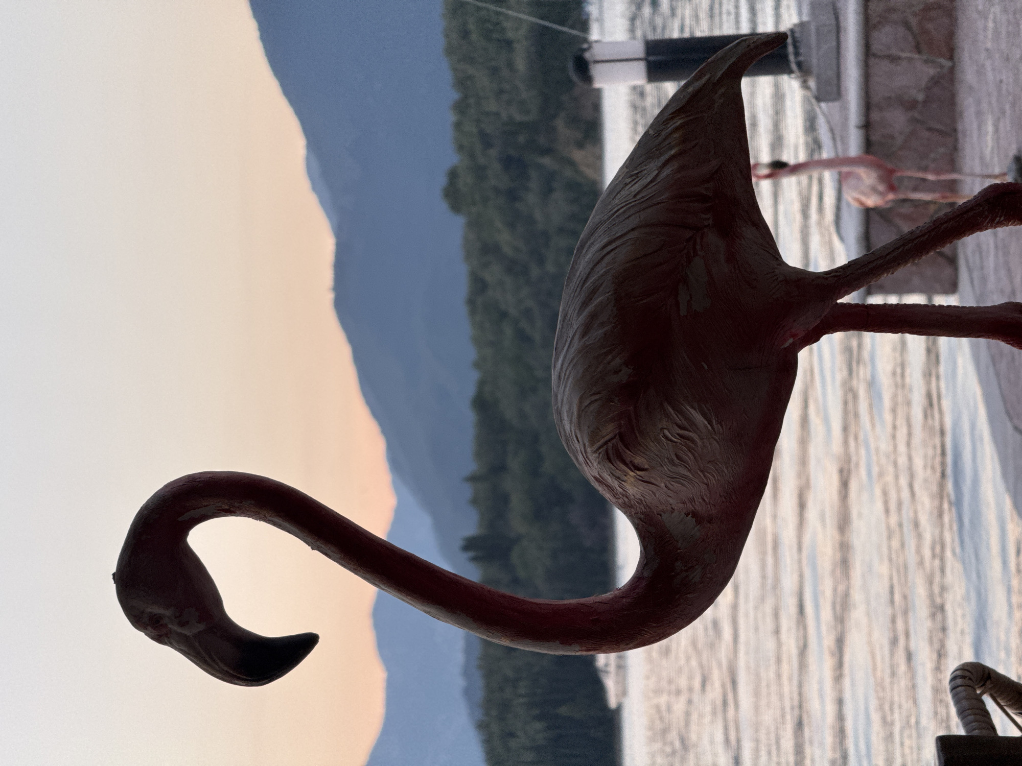 The image depicts a red flamingo statue in Bogišići, Montenegro. The statue is positioned near a body of water, possibly the bay, with mountains visible in the background under a pale sky. Another flamingo statue can be seen in the distance near a structure with wooden beams.