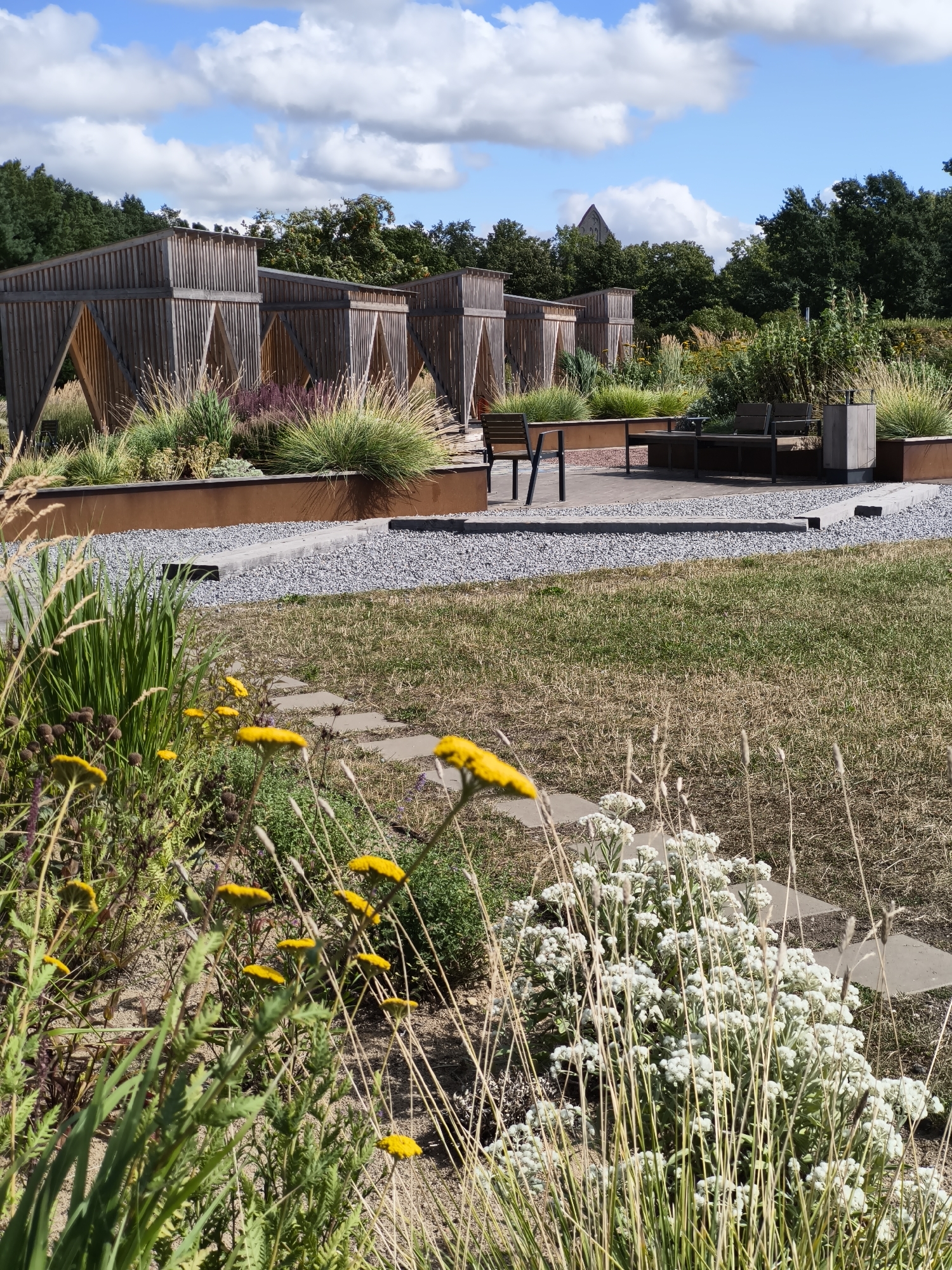 An outdoor scene features a modern garden design with geometric wooden structures in the background. In the foreground, a variety of plants and flowers are visible, including yellow yarrow and white blossoms. A stone pathway leads through a grassy area, and a seating area with benches and a table is set up on a gravel patio. The sky is blue with scattered white clouds. This garden is located at the Tallinn Botanic Garden in Tallinn, Estonia.