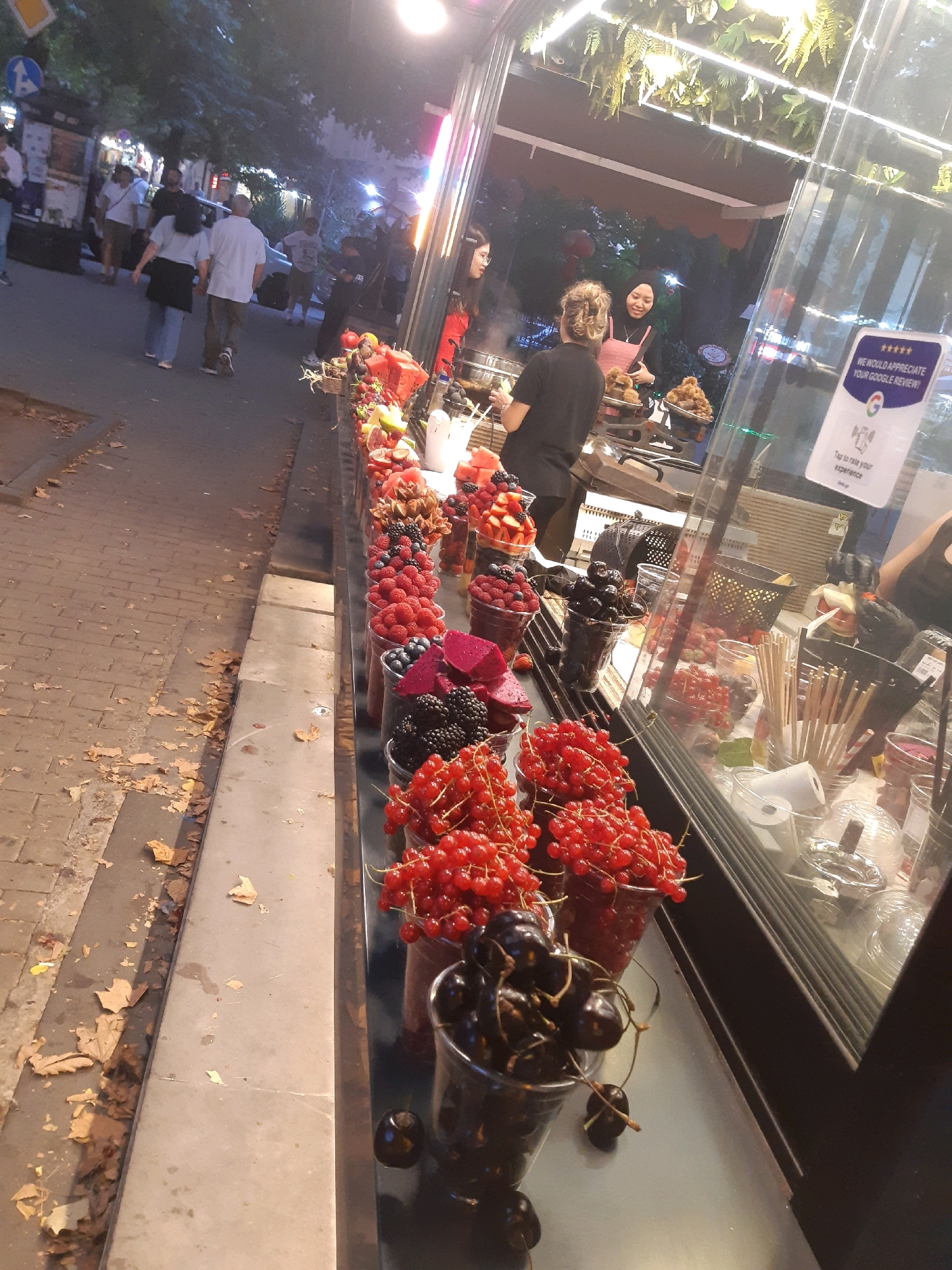 The image depicts a street scene in Tbilisi, Georgia, likely during the evening, given the artificial lighting. In the foreground, a glass display case is filled with various fresh berries and fruits, including red currants, blackberries, raspberries, blueberries, strawberries, and dragon fruit, arranged in clear plastic cups. Behind the fruit display, a glass-fronted establishment is visible, possibly a cafe or shop, with patrons and staff inside. One woman in a black shirt and another in a pink top are interacting within the establishment. Further in the background, pedestrians are seen walking on a paved sidewalk. The environment suggests an urban setting with trees and streetlights contributing to the ambiance.