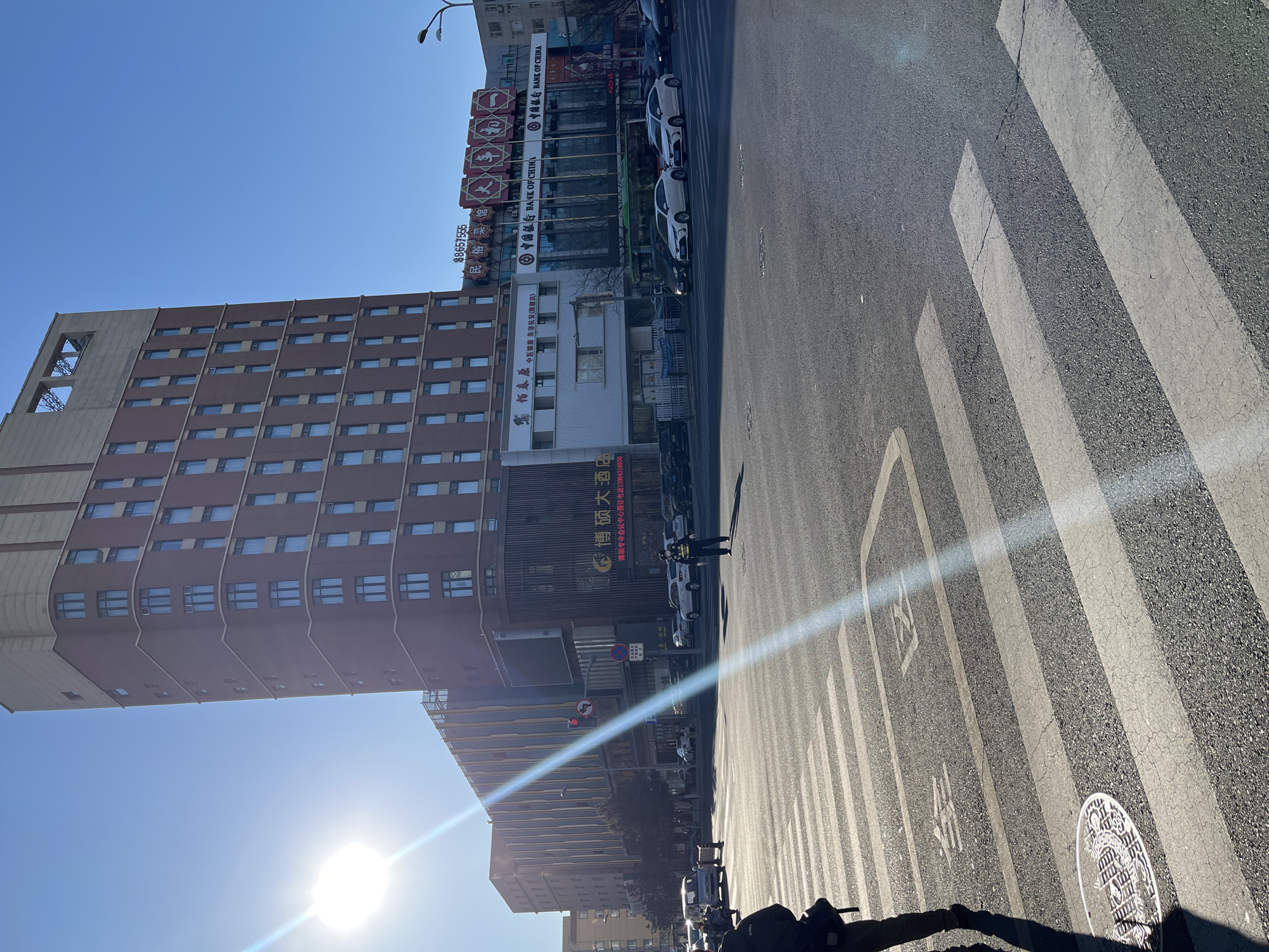 The image shows a street scene in Changchun City, China, under a clear, blue sky with the sun low in the sky. A tall, multi-story building, possibly a hotel or office building, dominates the left side of the frame, with numerous windows visible. Adjacent to it is another building with a sign in Chinese characters, advertising a hotel or restaurant named "博硕大酒店," along with a phone number.
The street is marked with a pedestrian crosswalk, and a traffic officer stands in the middle of the street, directing traffic. Several vehicles are parked along the right side of the road, including white cars. A Bank of China sign is visible on one of the buildings along the road. The street surface appears well-maintained. Markings on the street include the painted numbers 31 and 34. The overall environment suggests an urban setting on a sunny day.