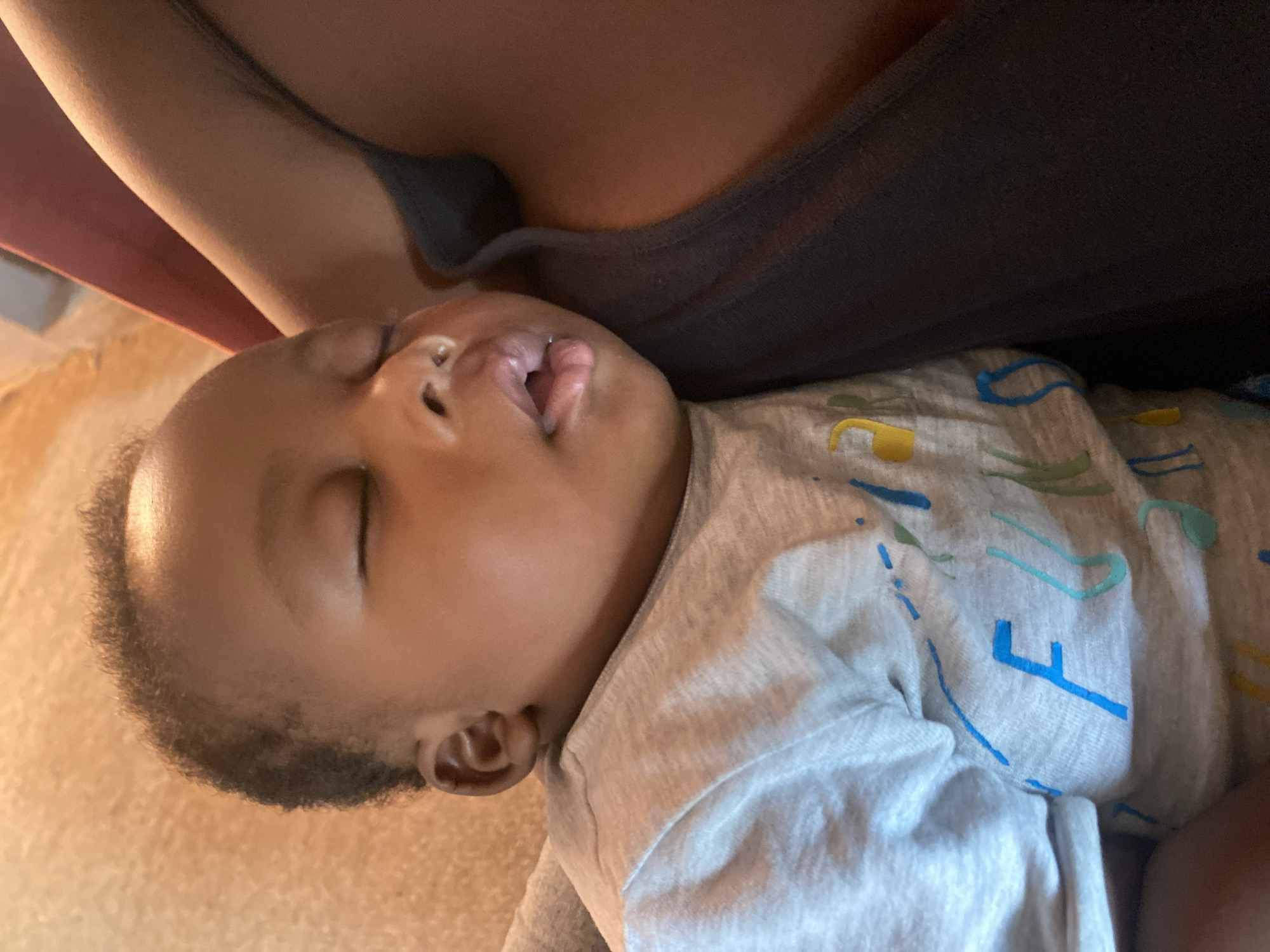 This image presents a close-up, intimate scene of a baby sleeping soundly, held or resting against an adult. The baby, with light brown skin and short dark hair, is positioned on their side with eyes closed and mouth slightly open, conveying a sense of deep relaxation and peace. They are wearing a light grey short-sleeved t-shirt featuring playful, abstract patterns in blue, green, and yellow hues.
In the upper part of the frame, a portion of an adult's shoulder and chest is visible, dressed in a dark grey or black garment, suggesting they are the one holding the baby. The background consists of a warm, light tan or beige surface, possibly a blanket, carpet, or furniture, contributing to the cozy ambiance.
The lighting is soft and warm, indicating an indoor setting, likely in the daytime or evening with artificial light. There are no explicit visual cues within the image to identify the specific location as Sabon Tasha, Nigeria, as provided in the context. The overall emotion evoked is one of tranquility and comfort. No discernible text is clearly visible on the baby's clothing or in the immediate surroundings; the patterns are purely abstract.