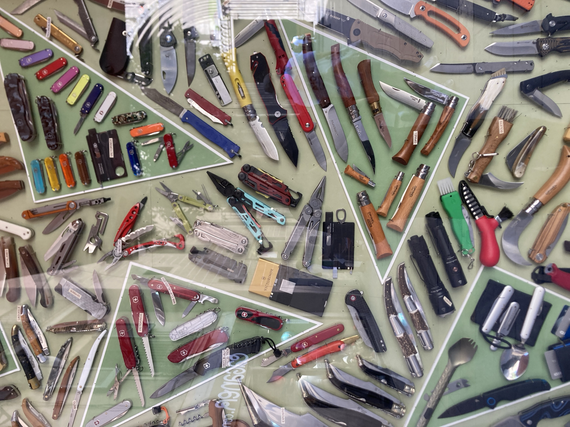 This image displays a large collection of knives and multi-tools, likely a retail display or personal collection, viewed through glass. The knives vary in size, color, and material, with some featuring wooden handles, polished metal, and bright plastic. Brands such as Victorinox, Opinel, and Nieto are visible on some of the items. The collection is arranged on a green background, possibly a felt or fabric surface, with some items laid flat and others displayed at an angle. Reflections from overhead lighting and the environment are visible on the glass surface, obscuring some details and making it difficult to ascertain specific contexts or actions. The image captures a diverse assortment of cutting implements, suggesting a focus on utility, craftsmanship, or collection. This display is located in Barcelona, Spain, inside a shop where knives are sold.