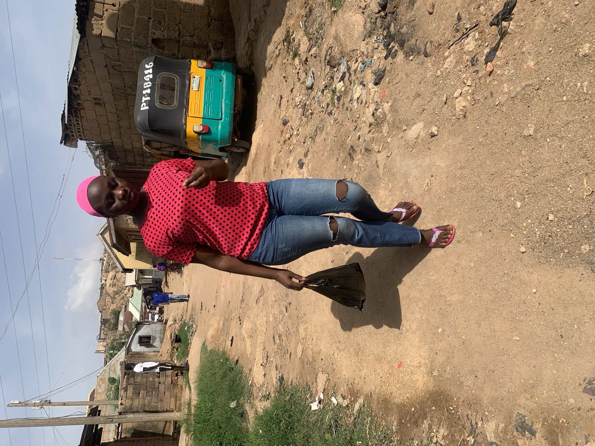 A medium shot captures a woman standing on a dirt road in Jos, Nigeria. She wears a red polka-dotted blouse, ripped blue jeans, and pink sandals, and her head is covered with pink fabric. She holds a black plastic bag in her left hand. A black, yellow, and green auto rickshaw with the inscription "PT-18416" is parked behind her to the left. The road is uneven and littered with debris. Buildings, some incomplete, line the street, and hills are visible in the distance. The sky is pale blue.