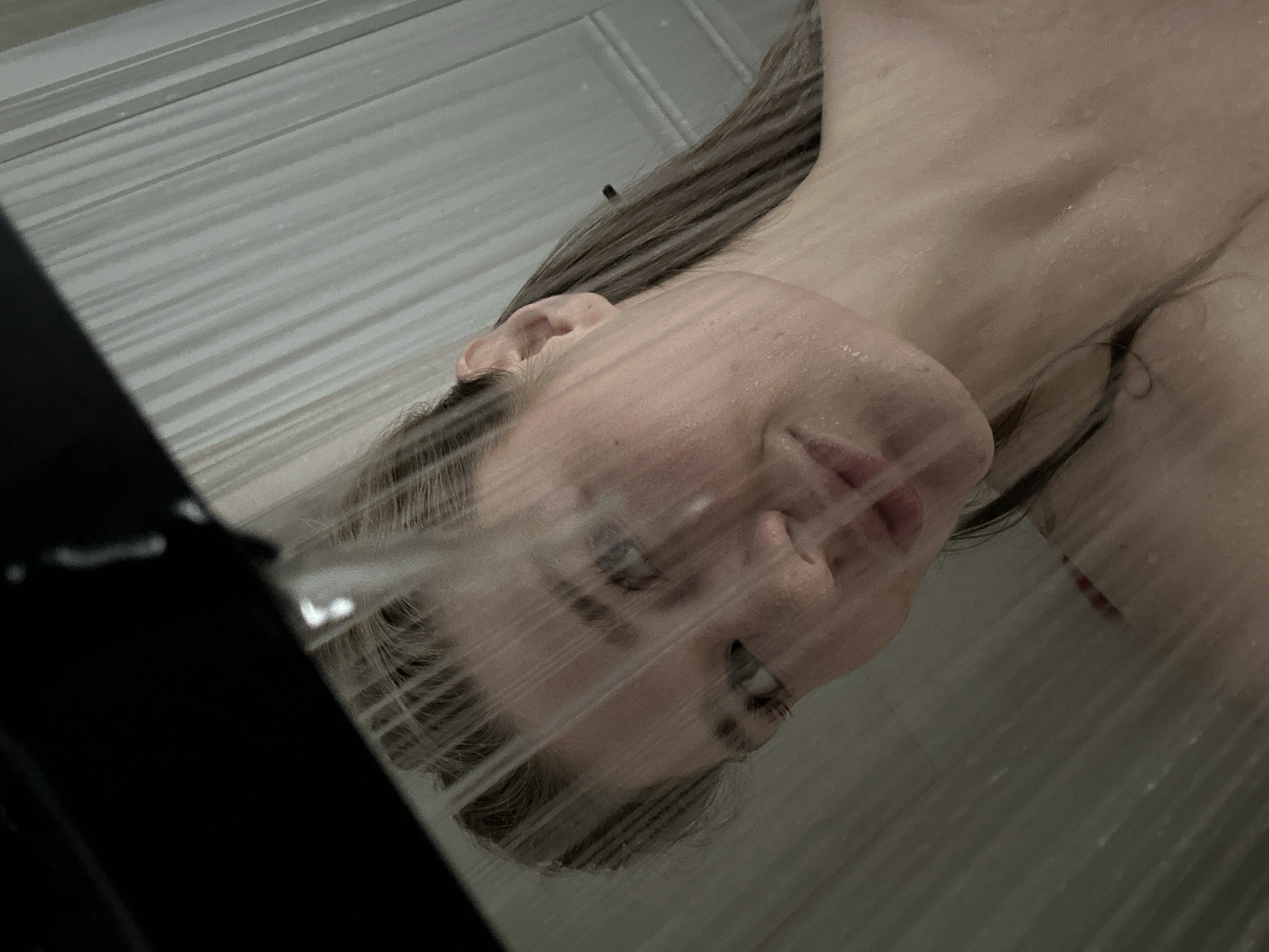 An indoor scene depicts a female individual showering. The subject, a woman with light brown hair, is positioned facing the camera, partially obscured by multiple vertical streams of water originating from an unseen overhead fixture, partially visible as a dark, angular object in the upper left foreground. Her face is wet, exhibiting water droplets, and she displays a slight, closed-mouth smile. Her gaze is directed towards the viewer. The visible background consists of a light-colored wall with vertical paneling or grooves, indicative of a shower enclosure. A faint red strap or garment edge is visible on her left shoulder. The specific location within the city of Kazan, Russia, cannot be determined from the provided visual information.