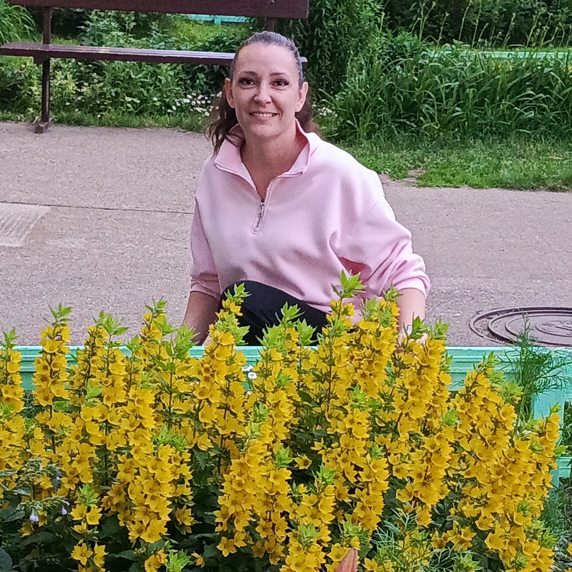 An outdoor medium shot captures a woman with dark hair pulled back and a friendly smile, wearing a pink zip-up sweatshirt and black pants. She is crouching in front of a vibrant display of bright yellow flowers. The background features a concrete path, a dark brown bench, and lush green foliage, suggesting a park or garden setting within the city of Markovsky, Russia.