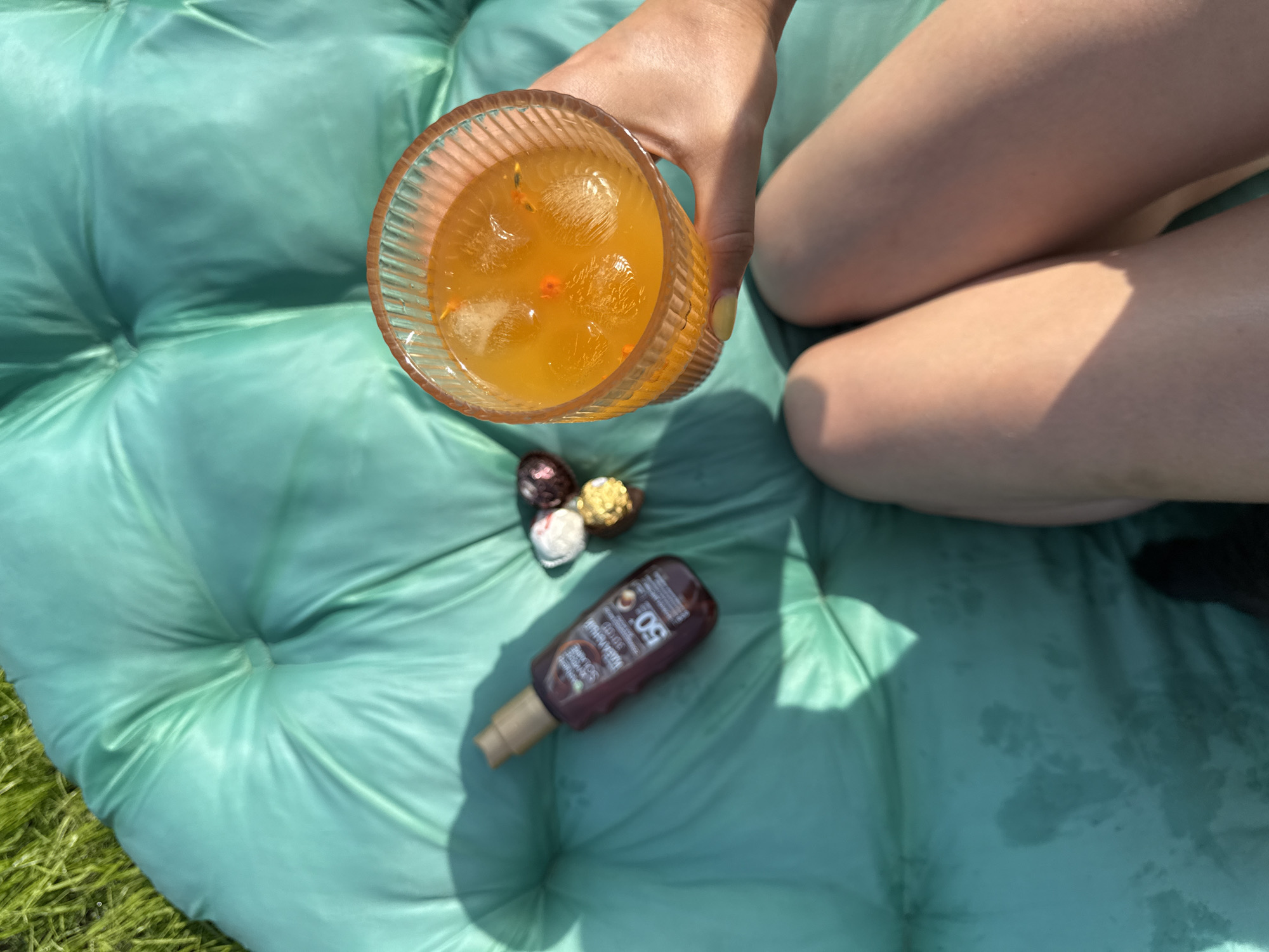 A person's legs are visible in the lower portion of the frame, resting on a teal inflatable mattress. The mattress is situated outdoors, possibly on grass, as indicated by the green blades visible in the upper left corner. The lighting suggests it is a sunny day, with shadows cast across the mattress.
On the mattress, to the left of the person's legs, is a bottle of sunscreen with SPF 50. Next to the sunscreen are three small, foil-wrapped chocolates. In the person's right hand, held towards the top right of the frame, is a glass of orange-colored beverage with ice cubes. The glass is ribbed and has a translucent orange tint.
The scene is likely taking place in a relaxed outdoor setting, perhaps a backyard or a park. While the image does not provide specific clues to pinpoint the exact location within Lobanovo, Russia, the presence of an inflatable mattress, sunscreen, and a refreshing drink suggests a leisure activity in a temperate outdoor environment.