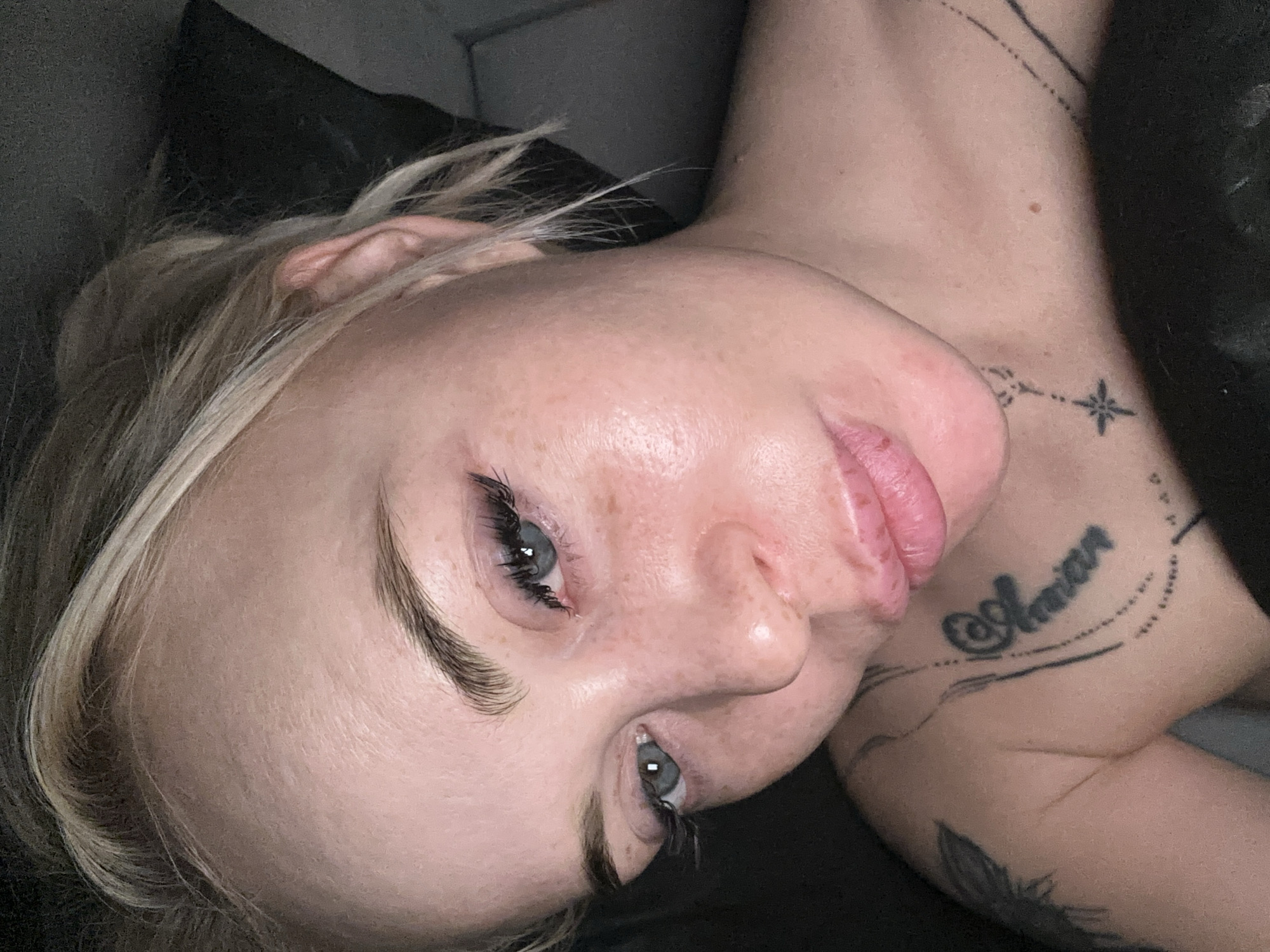 A close-up portrait shows a person, likely a woman, lying down with their head tilted. The person has blonde hair, which is partially visible around their ear and forehead, and light blue or grey eyes looking directly towards the viewer. Their skin shows some freckles across the nose and cheeks. Dark, well-defined eyebrows frame the eyes, which also feature long, dark eyelashes. The person's lips are slightly parted and appear full, with a pink hue.
The background is dark and indistinct, suggesting an indoor setting, possibly against a pillow or dark clothing. Several tattoos are visible on the person's neck and shoulder. On the right side of the neck, a delicate line of dots and dashes leads to a small star-like design. Below this, on the shoulder/upper chest area, a prominent script tattoo is visible, which appears to read "@amian" or a similar word, enclosed by curved lines above and below. Another faint line of tattooing is present further down on the arm. The overall mood conveyed is calm and relaxed. No specific time of day or location cues are identifiable beyond the indoor setting.
