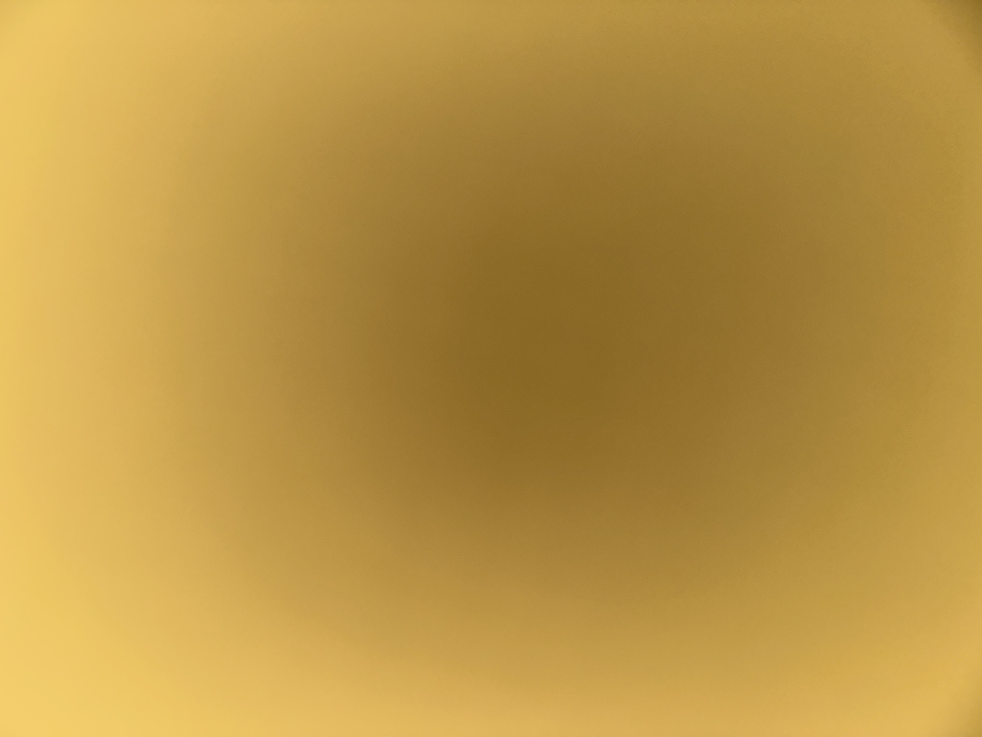 This media file is a close-up, out-of-focus abstract image with a gradient of golden-yellow hues. The color transitions from a brighter yellow on the left and bottom to a darker, browner-yellow tone in the upper right quadrant. The image appears to be a texture or surface with a slightly grainy quality, but no specific objects, people, or a discernible scene are visible due to the extreme blur and lack of detail. There is no activity or event taking place, and no notable details such as time of day, weather, location cues, or emotions can be identified. There is no visible text. The overall impression is that of a smooth, warm, and perhaps metallic or fabric-like surface, but without any clear context.