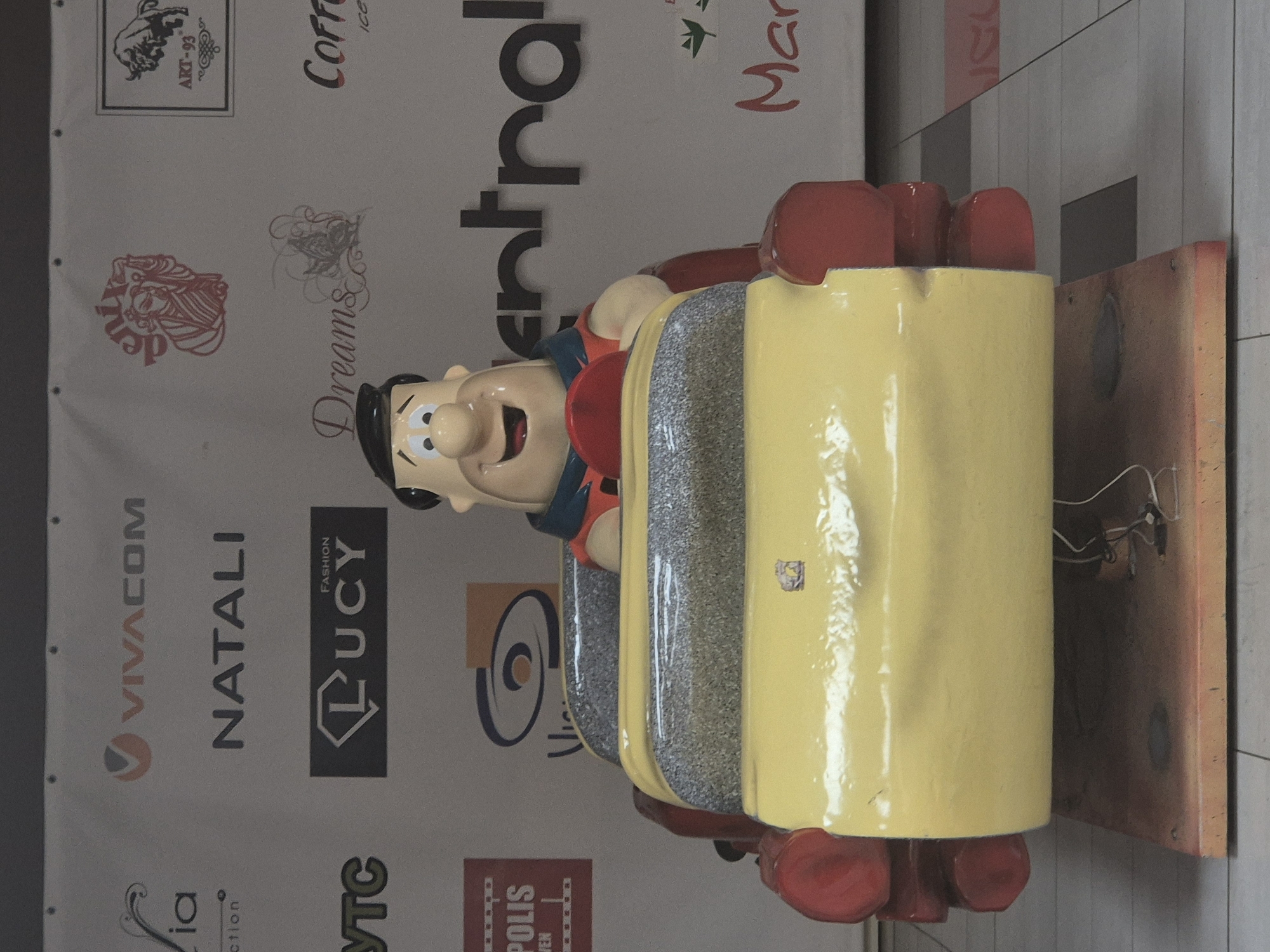A cartoonish figure of Fred Flintstone is seated in a yellow car, which is part of a children's amusement ride. The figure wears a red and blue outfit and has a wide smile. The car has a textured grey seat and red bumper-like elements on the sides. The background is a white banner with various logos and text, including "VIVACOM," "NATALI," "LUCY," "Dreams," "ART-93," "Coff," and "Mar." The ride is positioned on a wooden platform, which sits on a tiled floor. The overall scene appears to be indoors, possibly in a mall or entertainment venue.