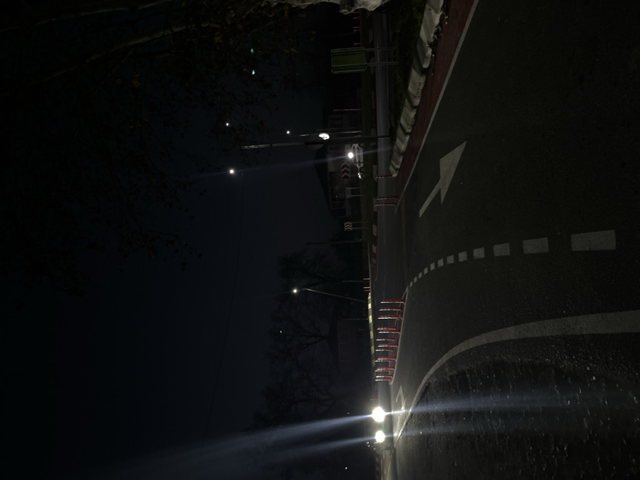 The image displays a nighttime scene of a road junction, specifically a roundabout, located in ლესის თემი, Georgia. The dark asphalt road curves through the foreground, featuring a solid white line along its inner edge and a broken white line separating a lane on the left. This lane is further defined by a series of approximately fifteen orange and white traffic delineator posts. A large white arrow is painted on the asphalt, indicating a right turn. The road's right edge features a raised concrete curb with a red-painted top surface.
On the left, a vehicle approaches with bright headlights, casting prominent, elongated beams of light onto the road surface. A second, lighter-colored vehicle with illuminated headlights is partially visible on the far side of the roundabout. Multiple streetlights provide artificial illumination across the scene, appearing as distinct light sources and glow points. In the background, several dimly lit buildings, including a two-story structure, are visible amidst what appear to be bare deciduous trees. Two red-and-white chevron signs, indicating a left curve, are positioned on the far side of the roundabout. A tree with dark foliage is present on the far right edge of the frame. The overall environment is characterized by low natural light conditions, with visibility primarily maintained by the artificial light sources and vehicle headlights.