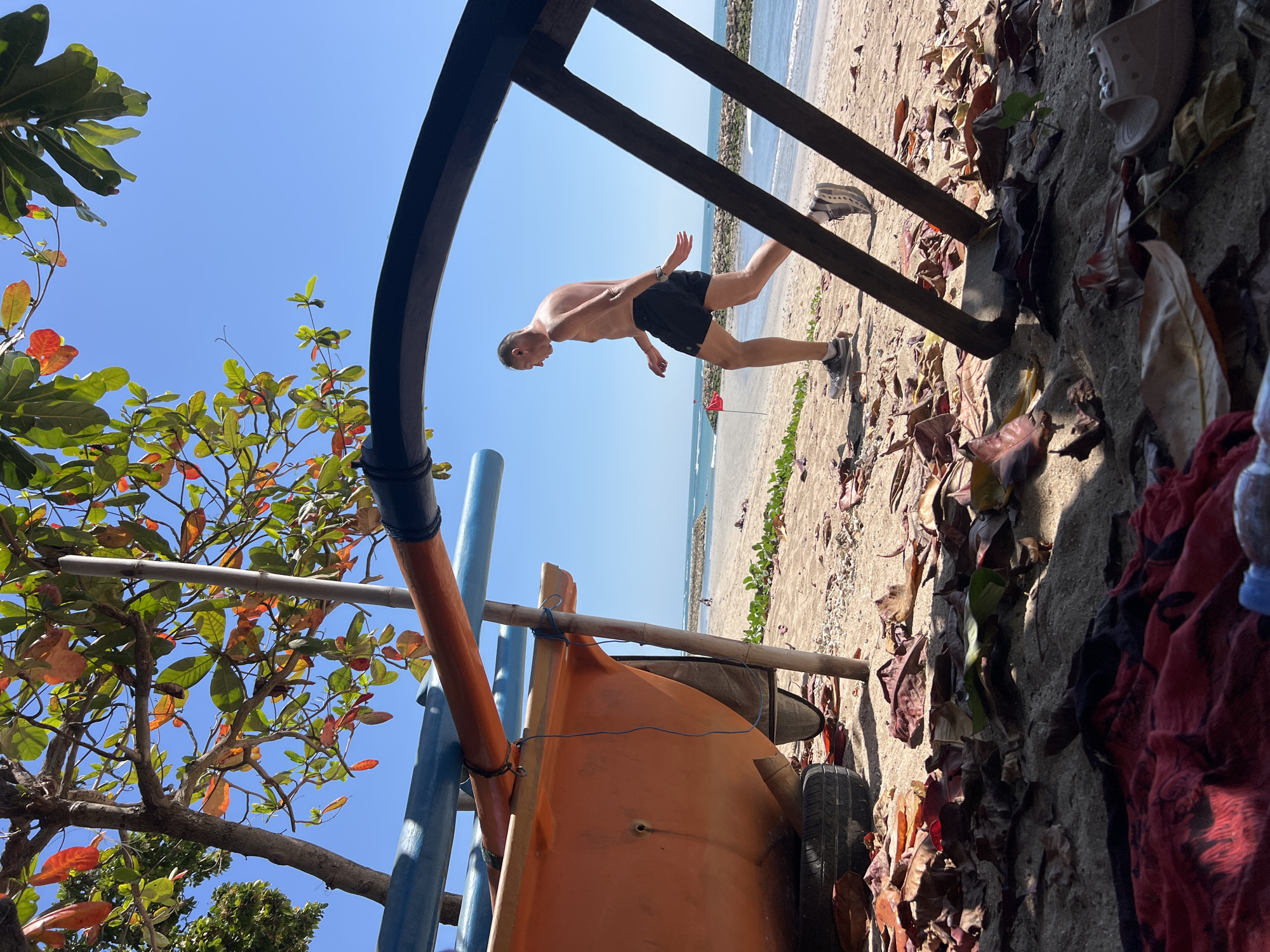 An adult male, wearing black shorts and grey athletic shoes, jogs on a sandy beach in Kuta, Indonesia, under a clear blue sky. The man is shirtless and appears to be sweating. In the foreground, a partially visible orange boat rests on the sand, supported by bamboo poles and a tire. The beach is scattered with fallen leaves and other debris. The ocean is visible in the background, with gentle waves and a rocky breakwater. The scene is framed by the branches of a tree with green and reddish leaves, as well as a dark, curved structure. The overall impression is of a casual, sunny day at the beach.