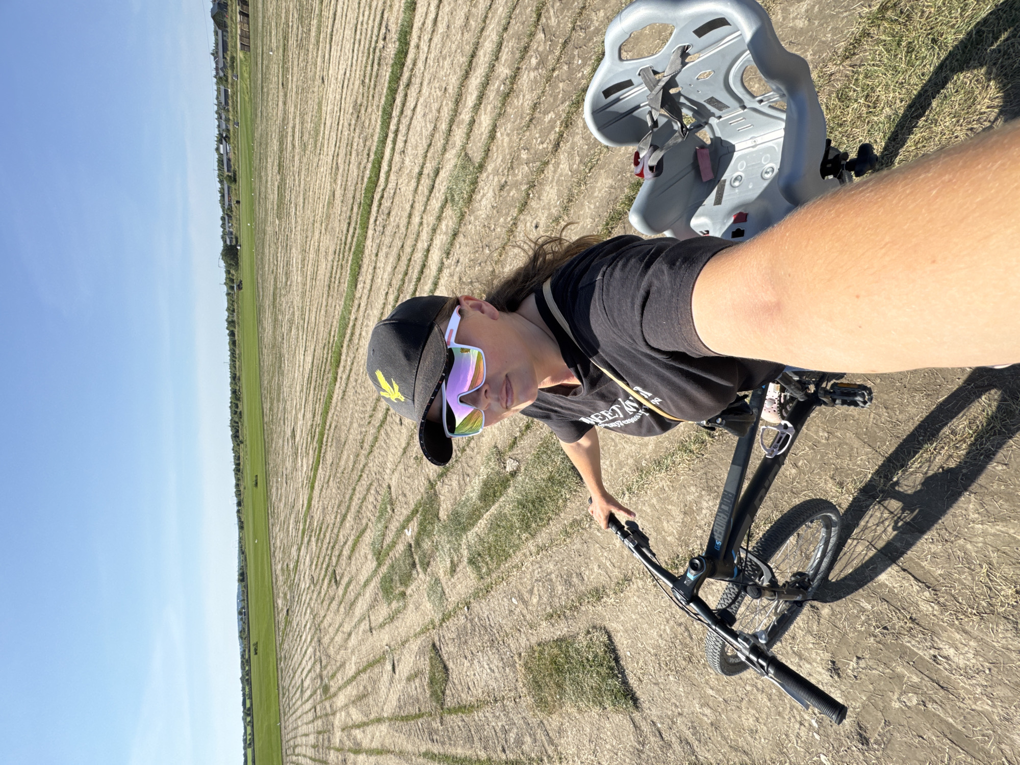 A woman, wearing sunglasses and a black baseball cap, is taking a selfie while riding a bicycle with a child seat. She is in an open field with rows of harvested grass or sod. In the background, there is a green field and some buildings. The sky is clear and blue. The scene appears to be in the city of Kempelevo, Russia.