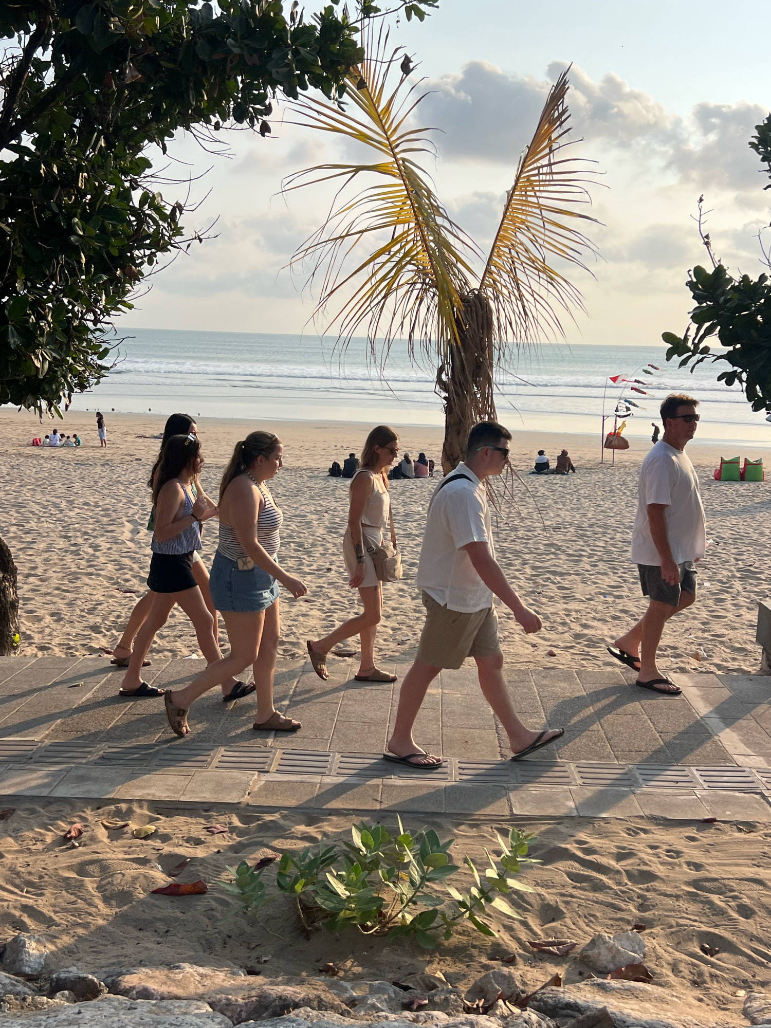 In Kuta, Indonesia, a group of people walk along a paved path bordering a sandy beach. The ocean stretches out in the background under a sky with scattered clouds. A lone palm tree stands prominently on the beach. Several individuals, some in casual attire such as t-shirts, shorts, and flip-flops, are depicted in motion. Others are seated on the sand in the distance. The scene is illuminated by natural sunlight, casting shadows on the path and sand.