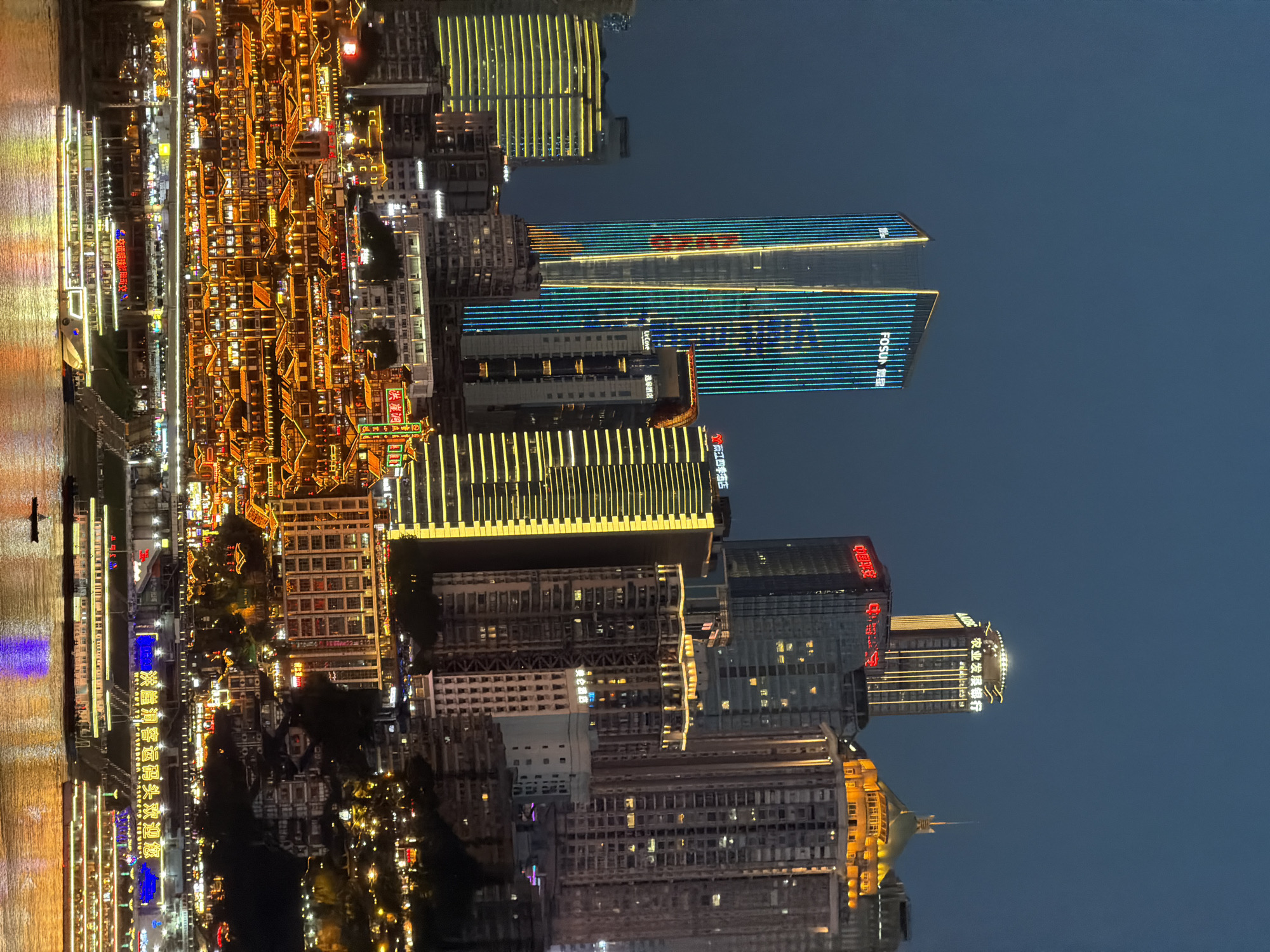 The image depicts a nighttime cityscape of Jiangbei Cheng Street, Chongqing, China, characterized by a dramatic juxtaposition of modern skyscrapers and traditional architecture illuminated by vibrant lights. In the foreground, a wide river reflects the city's glow. Several large passenger boats with illuminated decks are docked or cruising on the water. The middle ground features a densely packed area of multi-story buildings with traditional Chinese architectural elements, their windows and roofs aglow with warm orange and yellow lights, creating a cascade of illumination up the hillside. Further back, imposing modern skyscrapers rise into the dark sky, some featuring vertical streaks of blue and green lights, while others are crowned with illuminated signs. One prominent skyscraper displays the "FOSUN" logo in white lettering against a backdrop of blue and green vertical lights. Other buildings show yellow and white light patterns. The overall scene conveys a dynamic and visually striking urban environment at night.