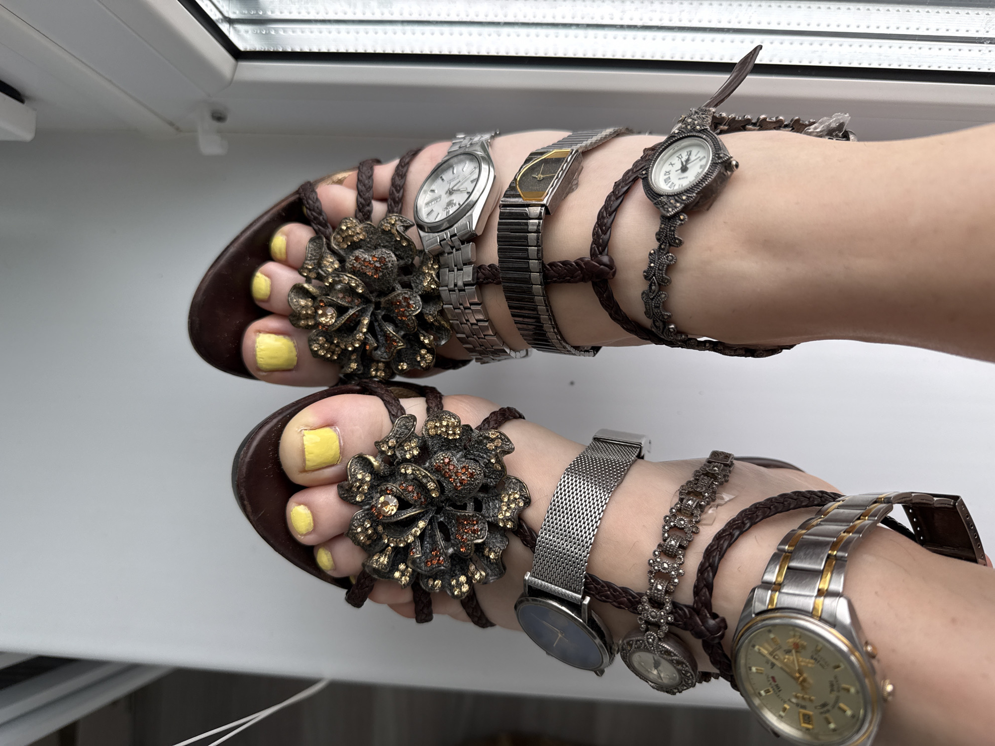 A close-up, overhead shot shows a pair of feet adorned with multiple wristwatches wrapped around the ankles and intertwined with the braided straps of dark brown, thong-style sandals. The sandals feature ornate, metallic floral embellishments on the toe straps. The toenails are painted a bright yellow. The feet are positioned on a white surface, with a blurred background suggesting a window with blinds or a pane of glass.