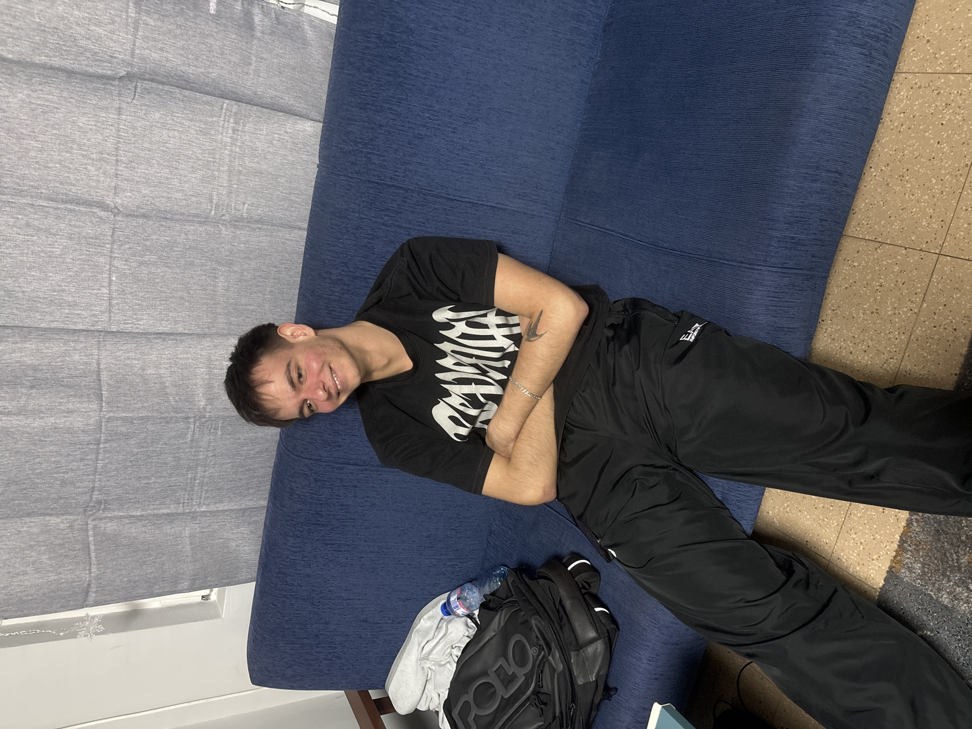 The image displays a young man lying horizontally across a textured blue sofa, smiling and looking directly at the camera. He has short dark hair and is wearing a black short-sleeved t-shirt with a white stylized graphic print that appears to say "Revenge" on the front. He is also wearing black athletic or track pants, with a small, partially visible logo that resembles "E-Sport" on the lower left leg. A silver chain bracelet is visible on his left wrist, and a small, dark tattoo is present on his right forearm.
Next to him on the sofa is a black backpack featuring the word "Polo" in grey letters. A folded light-colored garment and a clear plastic water bottle are placed on top of the backpack. The background features a light grey pleated curtain, likely covering a window, on the left. The floor visible on the right and bottom of the frame is a speckled pattern of beige, brown, and white tiles. The indoor setting appears to be a casual living space, well-lit, possibly during the daytime. The man's relaxed pose and smile convey a friendly and at-ease mood.