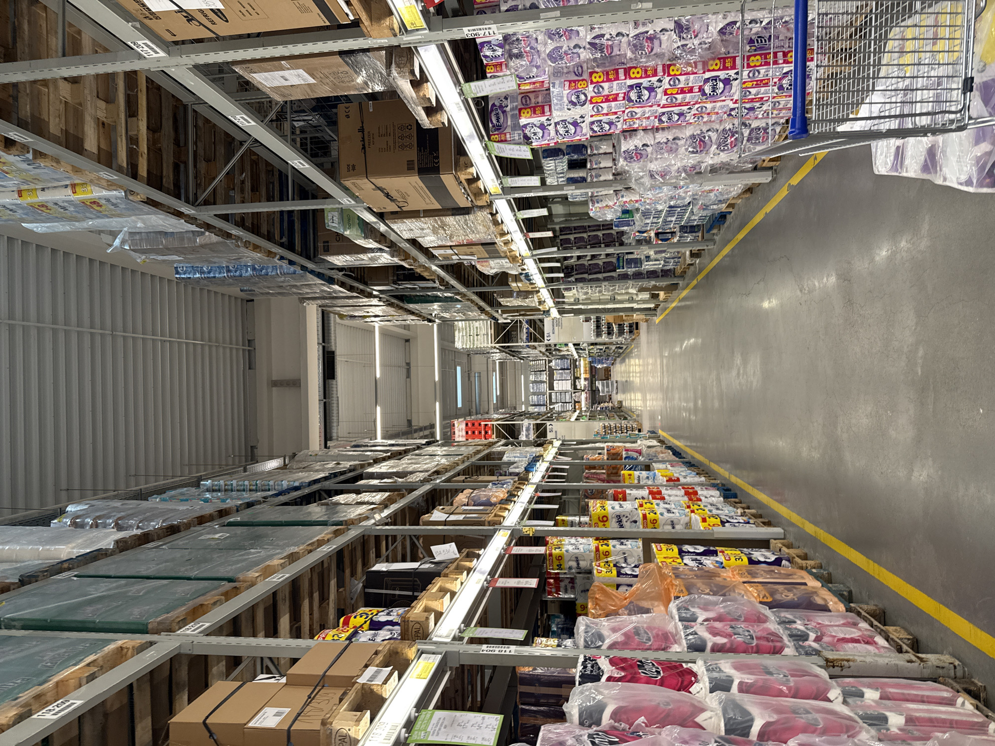 This is an interior shot of a large warehouse or wholesale store, potentially located in Stara Zagora, Bulgaria. The perspective is looking down a long aisle between tall shelves stocked with a variety of products. The shelves are metal racks holding wooden pallets loaded with goods. On the right, one can see items such as bottled water, toilet paper (branded "Emeka"), and packaged goods wrapped in plastic. There are also boxes with visible labels such as "FREEZER" and "REFRIG" indicating appliances or products needing refrigeration. The shelves on the left also display a variety of products. A yellow line runs along the floor of the aisle, marking the pathway for movement. In the upper-right corner, part of a shopping cart is visible, suggesting customers are present. The lighting is bright and artificial, common in large retail spaces. The overall impression is that of a well-stocked, industrial-scale storage or retail environment.