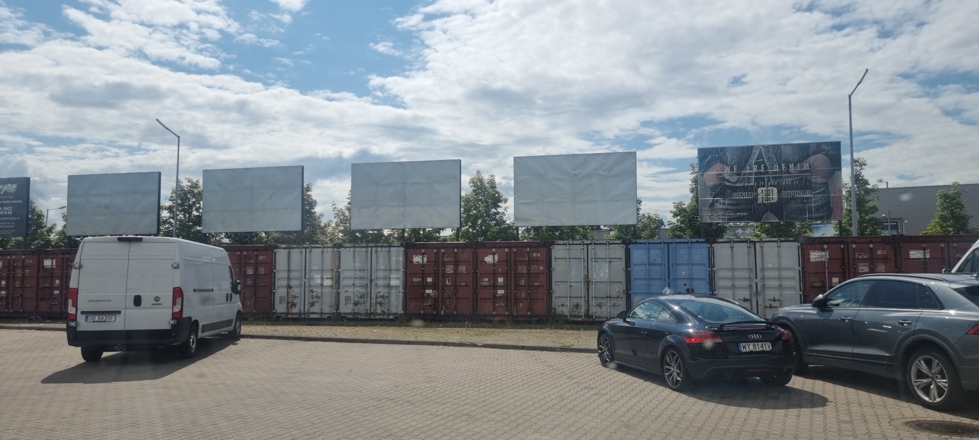 In Mroków, Poland, on a paved lot, a white van is parked facing away from the camera. To its right, a black Audi TT is parked. Further to the right, a grey Audi SUV is parked. Behind the vehicles, a line of shipping containers is visible, topped by several large billboards. The sky above is filled with scattered clouds.